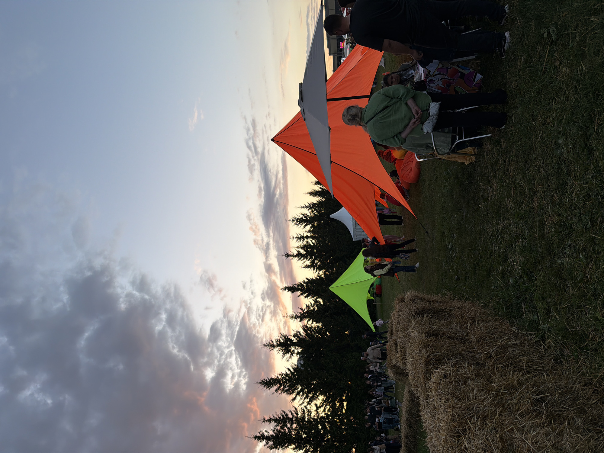 An outdoor event is taking place at dusk with a vibrant sky. In the foreground, several bales of hay are stacked. Beyond the hay, a crowd of people are gathered, some standing and some sitting. There are at least two colorful tents set up, one neon green and one bright orange, providing shade. An umbrella is positioned in front of the orange tent. In the background, a line of dark evergreen trees forms a silhouette against the sunset sky. The overall scene suggests a community gathering or festival.