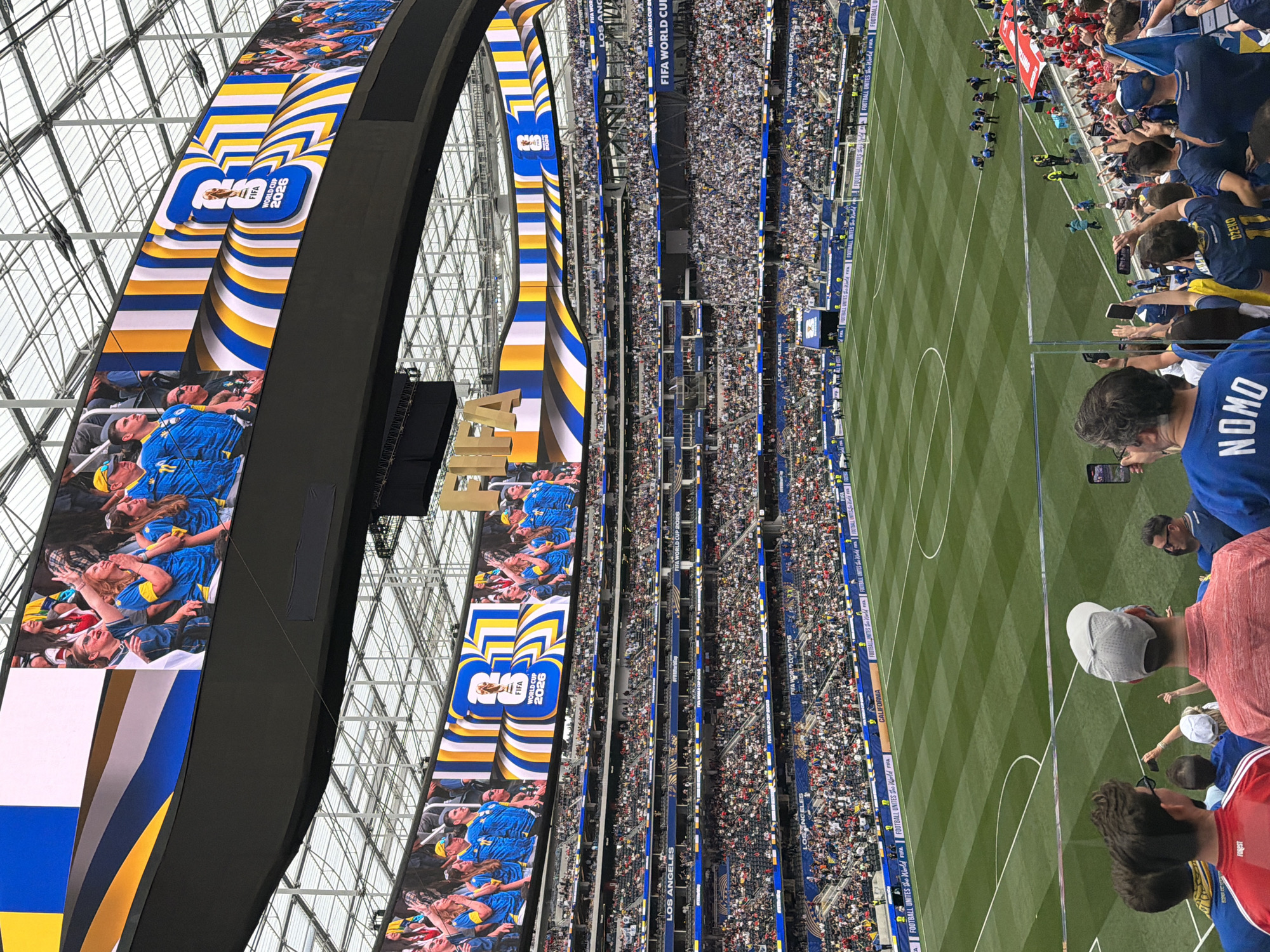 This is a wide-angle, elevated shot of the SoFi Stadium in Inglewood, California, USA, during a soccer match. The stadium is filled with spectators, and large screens display graphics related to the FIFA World Cup 2026. The field of play is visible in the foreground, with green grass marked by white lines. Several people are standing in the lower section of the stadium, some holding up their phones to take pictures or videos. The architecture of the stadium, including its retractable roof and tiered seating, is clearly visible. The overall atmosphere appears to be one of excitement and anticipation for a major sporting event.