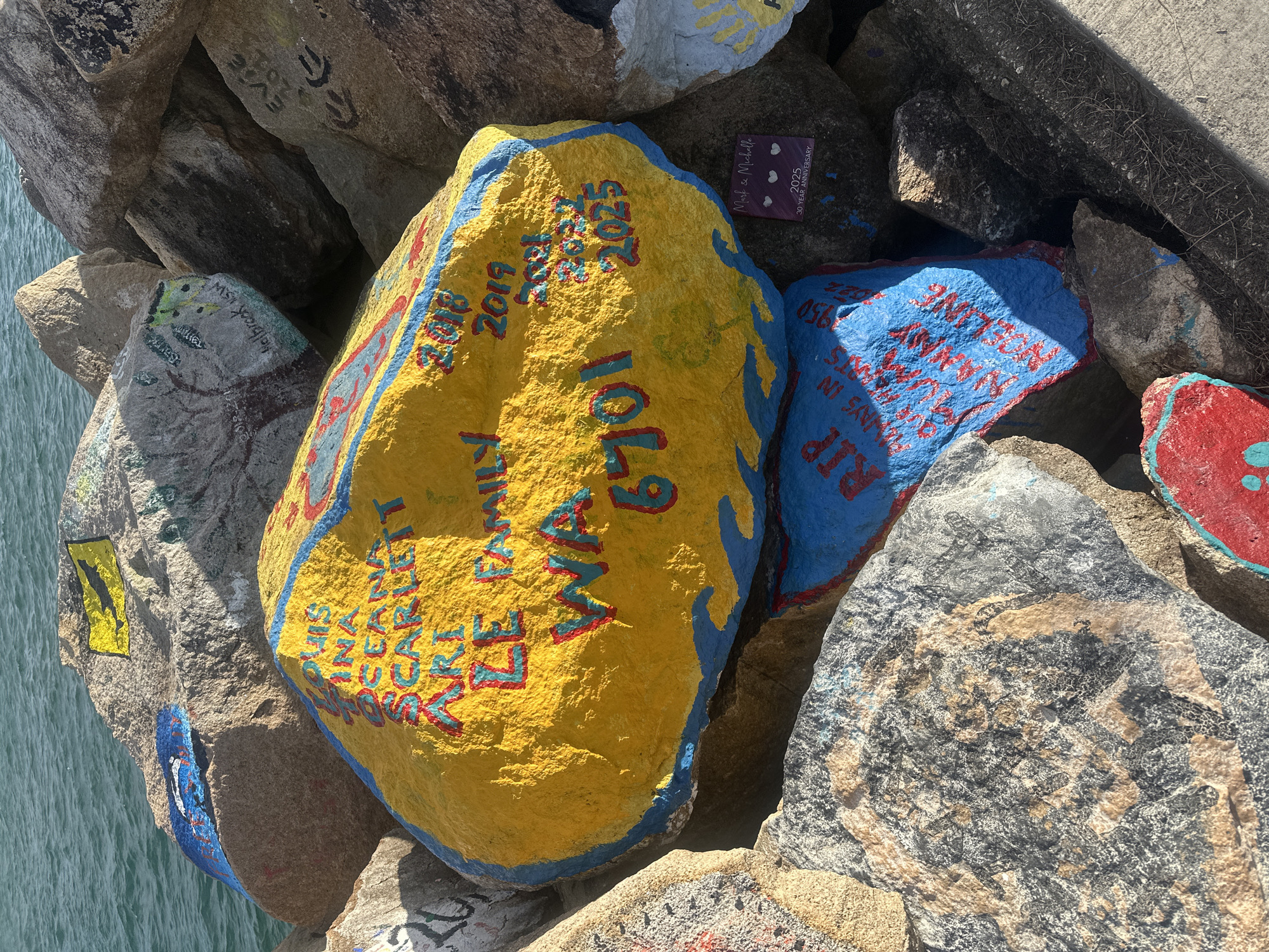 The image displays a collection of rocks painted with various messages and dates. The prominent rock in the center is painted yellow with a blue border and contains text in red and teal. Some of the visible text includes "THIS INA OCEAN SCARLETT ARI LE FAMILY WA 6701" and dates like "2018," "2019," "2021," "2022," and "2025." To the lower left of this central rock is another rock painted blue with red text, partially obscured, which appears to be a memorial with the word "RIP" visible. A small, square plaque is attached to a rock in the lower right, reading "Mark & Michelle 2025 30 YEAR ANNIVERSARY" with three heart icons. Other rocks in the background have faded or partial paintings, including a yellow flag with a black bird on one, and inscriptions like "Evie 2018" and "Hellbrook NSW" on others. The rocks are situated near a body of water, likely the sea, as indicated by the blue expanse in the upper left portion of the image. The overall scene suggests a public space where people have gathered to leave painted messages on rocks. This practice is commonly seen at landmarks like the V-Wall in Nambucca Heads, Australia, which serves as a memorial and a place for community expression.