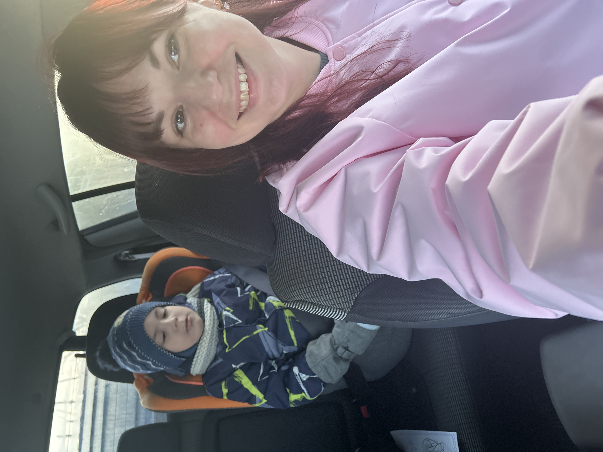 A woman with red hair and wearing a pink jacket is taking a selfie in a car. Behind her and to the left is a young child asleep in a car seat. The child is wearing a blue and gray jacket and a blue striped hat. The car is traveling in the city of Фоки, Russia.
