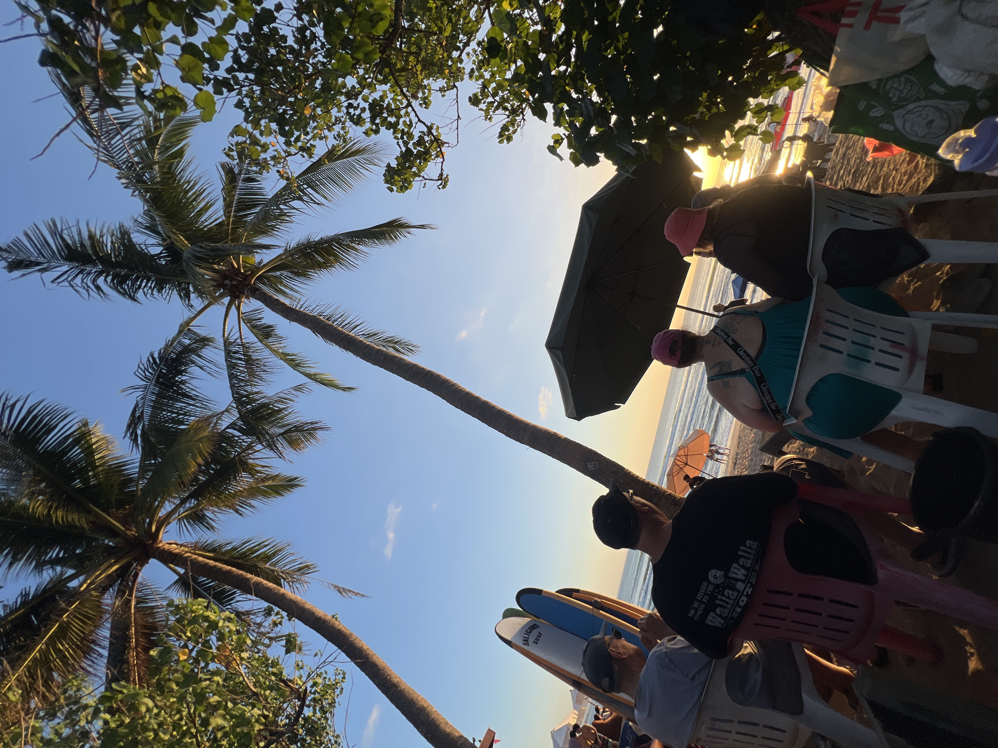 The image depicts a sunset scene on Kuta Beach, Bali, Indonesia. Two palm trees frame the shot, with their fronds silhouetted against a clear blue sky transitioning to a warm orange hue near the horizon. In the foreground, four people are seated on plastic chairs on the sand, facing the ocean. The individuals are observed from behind. The person on the far left wears a gray t-shirt and a baseball cap. Next to them, a person in a black t-shirt with white text is visible. The third person from the left is wearing a teal dress and a pink cap, and the fourth person wears a dark jacket and a red hat. They are seated under a large, dark umbrella.
In the mid-ground, several surfboards stand upright, leaning against a railing. The ocean stretches out to the horizon, with gentle waves breaking on the shore. A few other beachgoers can be seen in the distance, along with colorful beach umbrellas and tents. The sand is a light brown, characteristic of the beach. The overall atmosphere is relaxed and serene, capturing a typical evening on this popular Indonesian beach.