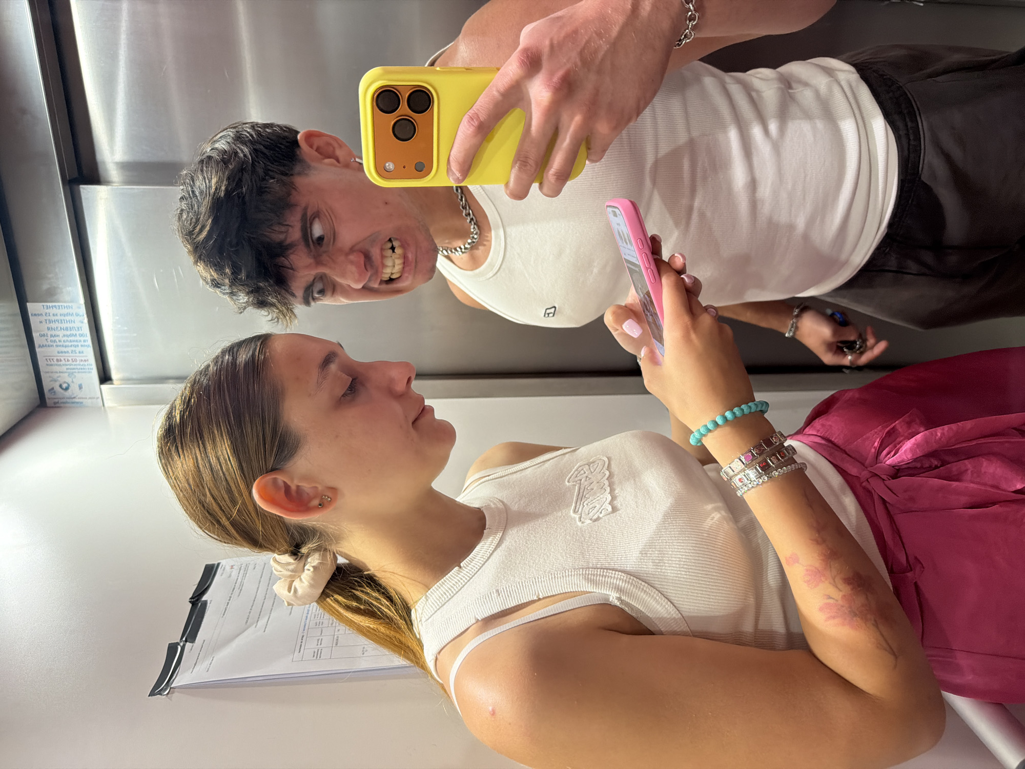 The image depicts two individuals in what appears to be an elevator in Sofia, Bulgaria. On the left, a young woman with her hair pulled back in a ponytail is wearing a white tank top and a bright pink skirt. She is looking down at a pink smartphone she is holding in her hands. On her left arm, she has a floral tattoo and is wearing several bracelets, including one made of turquoise beads.
To the right, a young man with dark, styled hair is making a distorted, angry facial expression directly at the camera. He is wearing a white tank top and dark sweatpants, and a silver chain around his neck. He is holding a yellow-cased smartphone in his right hand, also pointed towards the camera, as if taking a selfie with the woman. The elevator's stainless steel doors are visible in the background, along with a poster or document attached to the wall on the left side. The overall lighting suggests an indoor, possibly artificial, environment.