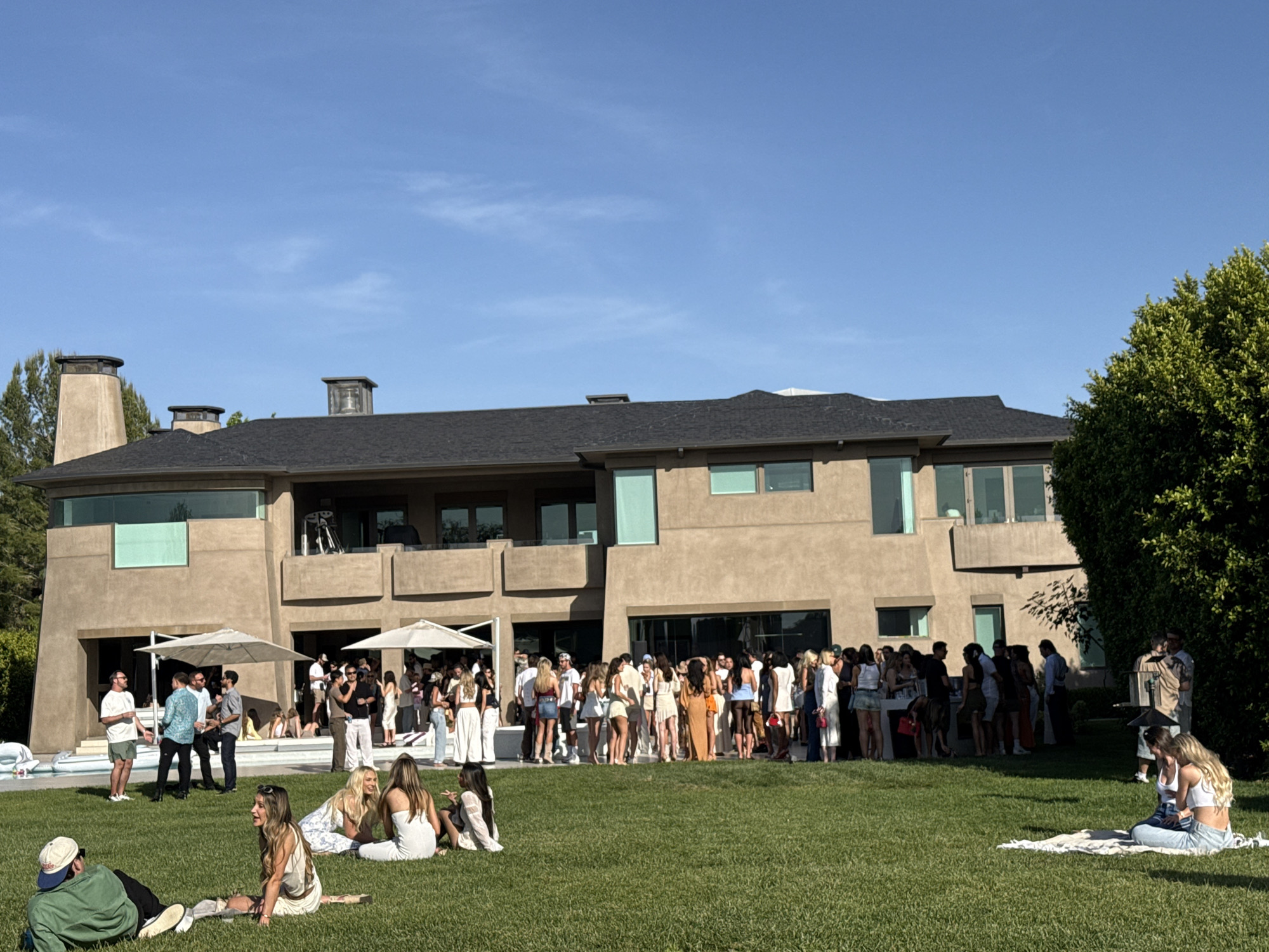 The image captures a large outdoor gathering, possibly a party, on a well-manicured lawn in front of a modern, two-story house. Approximately fifty or more people are present, most of whom are standing in a large group closer to the building, while others are sitting or standing in smaller groups on the lawn. The attire of the people is generally casual and summery. The building is light brown with a dark roof and large windows. There are two white umbrellas on the left side of the building, near what appears to be a pool area. The scene suggests a warm, sunny day with a clear blue sky. The location, based on architectural style and vegetation, could be a residential area in Los Angeles, United States of America.