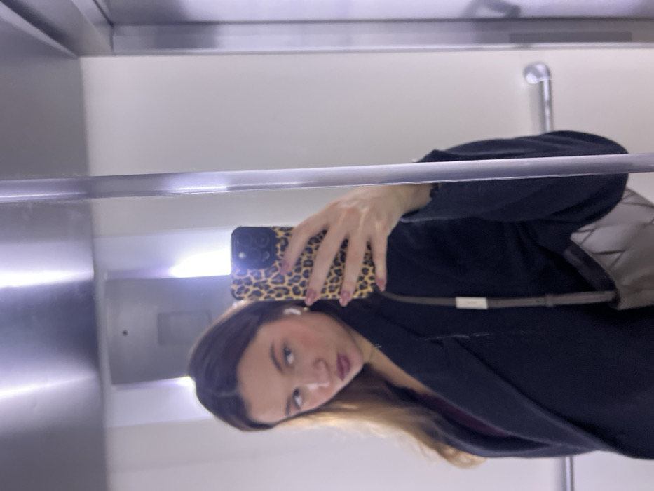 A woman takes a selfie in a reflective elevator. She wears a dark jacket and carries a patterned phone case. Her hair is partially visible. The elevator walls are plain and there is a silver handrail. A portion of a bag strap is seen.