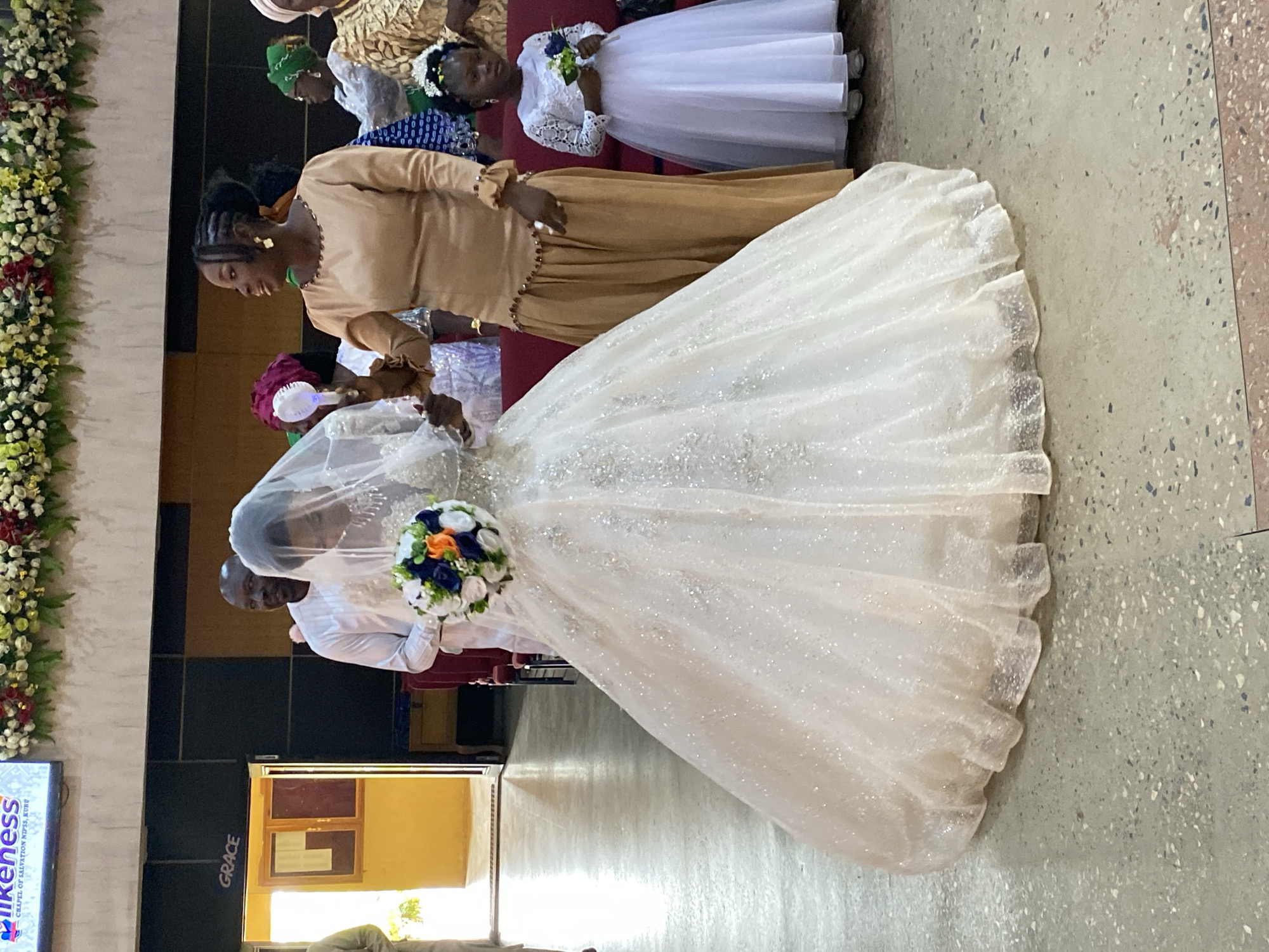 A wedding ceremony is taking place in Kuru Karama, Nigeria. The bride, adorned in a sparkling white ball gown and veil, walks with a bouquet of blue, orange, and white flowers. She is accompanied by a man in a white outfit and a woman in a tan dress holding a fan. To the right, a young flower girl in a white dress stands with a small bouquet. The ceremony appears to be held in a church, with floral decorations visible at the top of the frame. The floor is a speckled concrete. A sign in the background reads "GRACE".