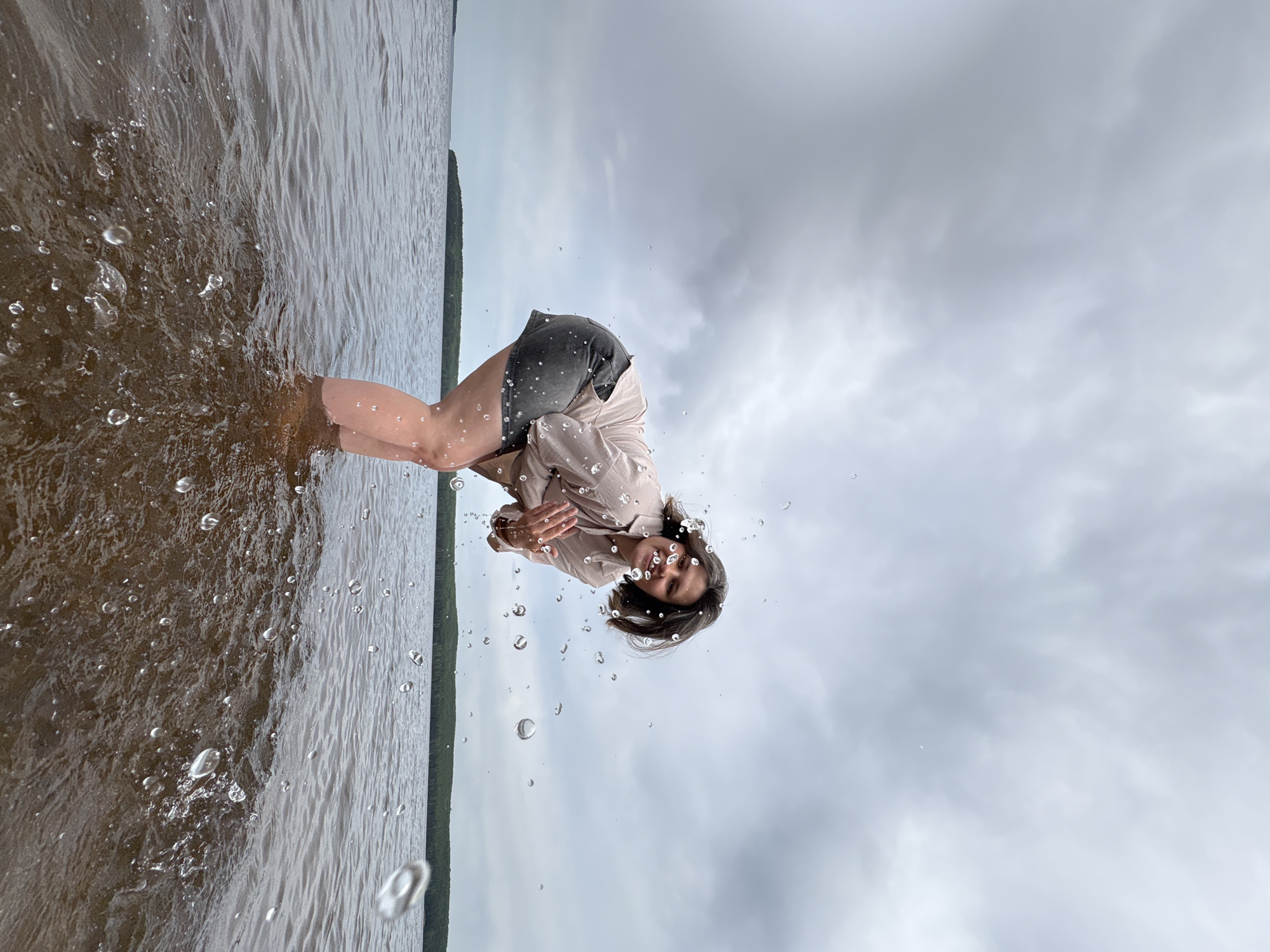 The image shows a person, likely a young woman, bending over in the shallow water of a lake or river. She is wearing shorts and a light-colored shirt. The water is murky, and there are splashes around her. The background shows an overcast sky and a line of trees on the opposite shore. The location is Chaikovsky, Russia, based on the subject's presence there. The person is likely playing or interacting with the water.