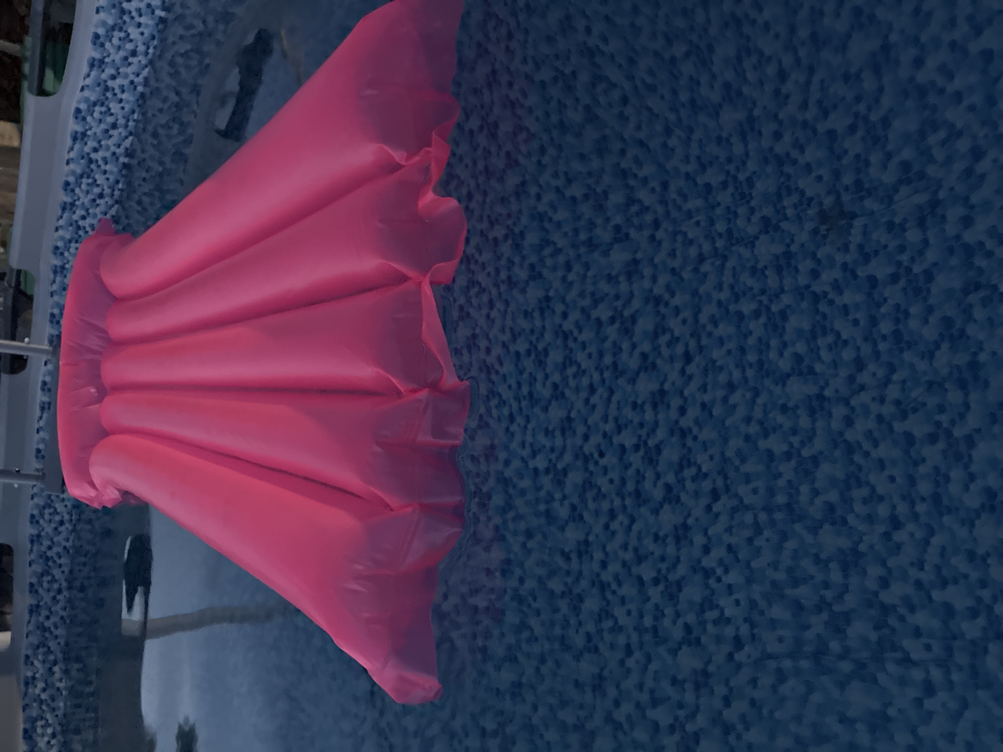 The image shows a bright pink inflatable pool float in a swimming pool. The pool water is a deep blue and the pool liner has a speckled blue and white pattern. The float is partially submerged in the water and its inflated sections create a ribbed, fan-like shape. The top edge of the pool and some railings are visible in the background. The scene appears to be outdoors, with daylight reflecting off the water. The specific location of Totskoye, Russia, cannot be determined from the visual information in the image.