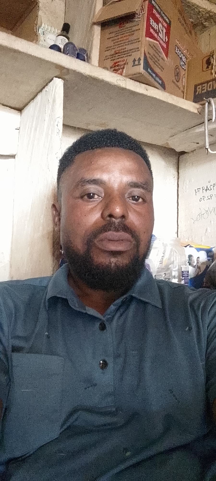A man with dark skin, a beard, and short hair is shown in a close-up, waist-up shot. He is wearing a dark teal collared shirt with two dark buttons visible. The background appears to be an indoor setting with shelves. On the shelf behind him, there are boxes and some bottles. One box has red and white lettering and a "15 FREE" advertisement. To the right of the man, on a wall, there is some writing that appears to be "PASS OP SP". The lighting is somewhat dim, with a soft glow illuminating the man's face. The overall scene suggests a casual, perhaps personal, setting.