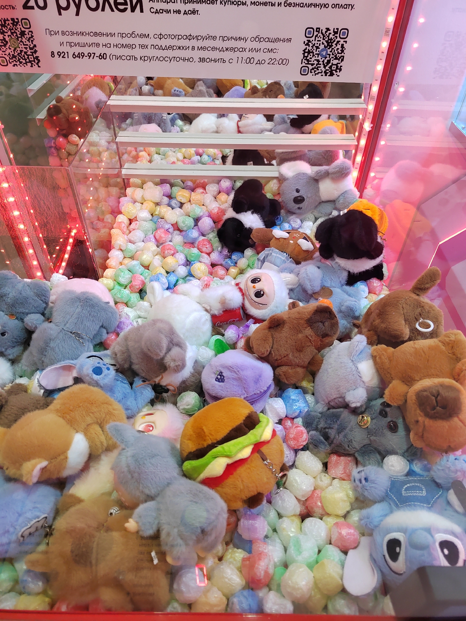 The image depicts the interior of a claw machine filled with an assortment of plush toys and colorful, individually wrapped prizes. The prizes appear to be small, spherical, and brightly colored in shades of pink, yellow, green, blue, and white, possibly bubble wrap or candy. The plush toys are diverse, including animals like dogs, cats, and possibly a koala or bear, as well as cartoon characters. Some of the plush toys are partially obscured by the prizes. The claw mechanism of the machine is visible at the top of the image. A sign in Russian is affixed to the top of the machine, with text that includes currency denominations and contact information. This claw machine is located in a public space, likely a shopping mall or entertainment center, in the city of Novogorelovo, Russia. The lighting is bright and likely artificial, with pink and red hues reflecting off the glass and toys, suggesting an indoor environment. There are no people visible in the immediate scene, but the presence of the machine implies a commercial or recreational setting where people would be interacting with it.