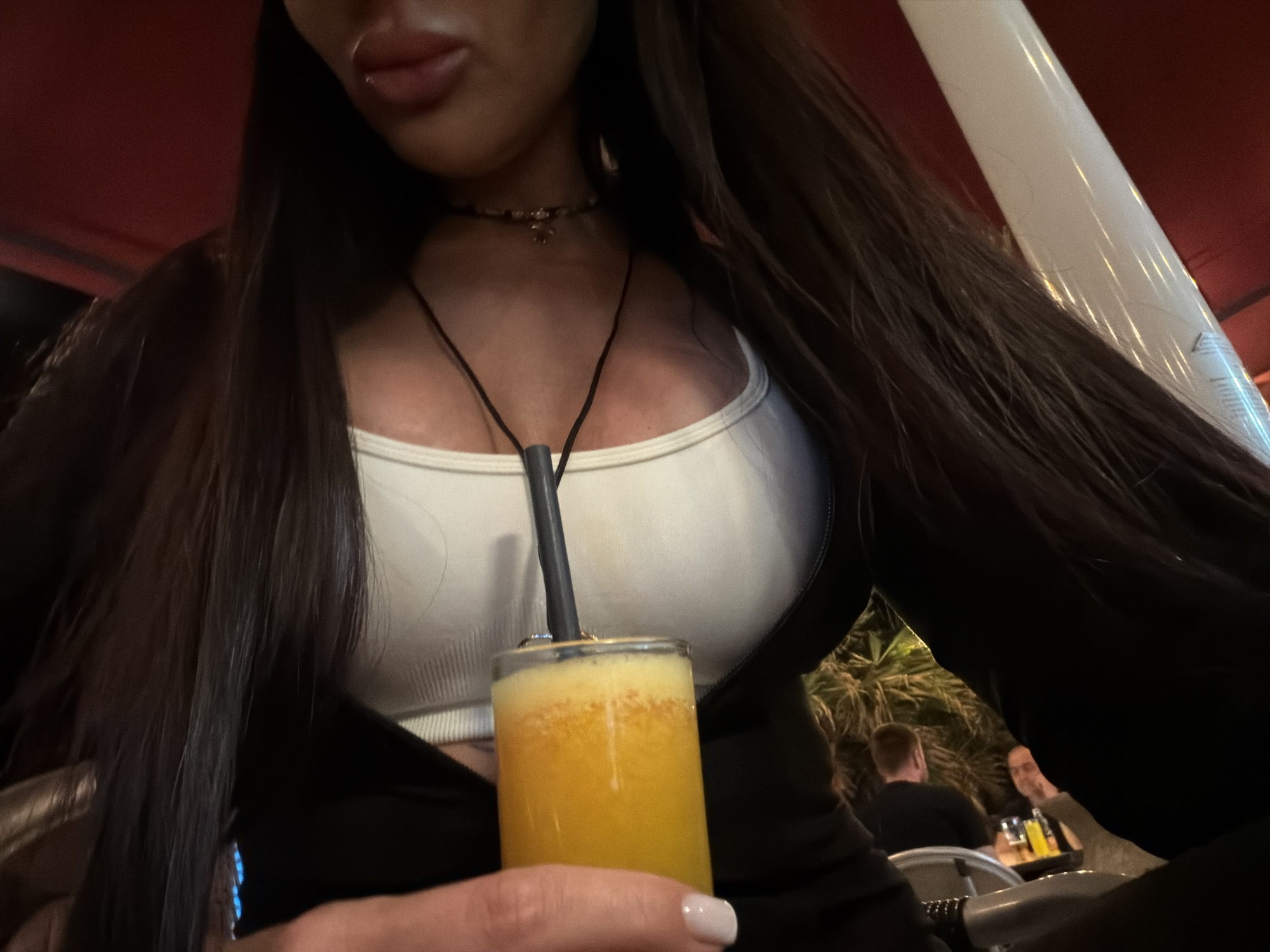 The image captures a partial view of a woman holding a glass of a frothy, orange-colored drink with a dark straw. She is seen from the chest up, with her long, dark hair flowing over her shoulders. Her face is mostly out of frame, but her full lips, possibly with a piercing, and chin are visible. She is wearing a white crop top or bralette underneath a dark open jacket or cardigan. A delicate choker necklace adorns her neck, along with a longer dark cord necklace. Her left hand, featuring a thumb with a white polished nail, holds the glass.
The setting appears to be an evening or night scene, likely at an outdoor or semi-outdoor cafe or bar in Rafailovići, Montenegro, given the dim lighting and reflections. In the blurred background, two men are seated at a table. One is seen from behind, while the other is partially visible, smiling towards the camera. Dark foliage, possibly palm trees, can be seen behind them, and bottles are present on their table. A white pole or pillar is visible on the right side of the frame, contrasting with a reddish ceiling or awning above. The overall activity suggests a relaxed, social gathering. No discernible text is visible in the image.