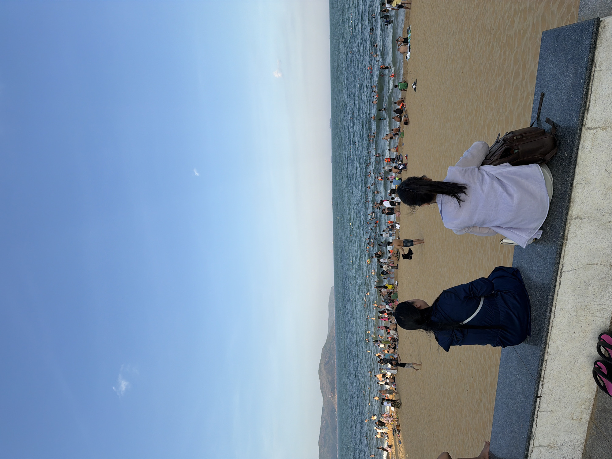 The image depicts a crowded beach scene in South Quy Nhon Ward, Vietnam. The foreground shows two women sitting on a concrete ledge, facing away from the camera and towards the sea. The woman on the left wears a dark blue hooded jacket. The woman on the right wears a light purple long-sleeved shirt and has a brown backpack beside her. In the background, a wide expanse of sandy beach is filled with numerous people swimming, sunbathing, and walking. The sea is populated with many individuals enjoying the water, and several boats or ships are visible on the horizon. In the distance, a mountain range borders the coastline under a clear blue sky with a few wispy clouds. The overall impression is a vibrant and active beach day.