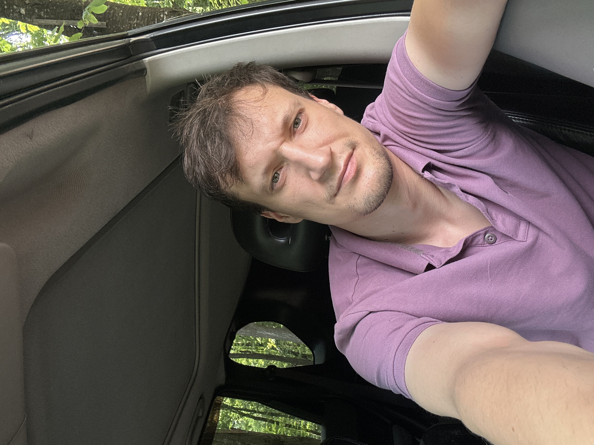 A man is taking a selfie inside a car. He is wearing a purple polo shirt and has short brown hair. The car appears to be parked on a dirt road or in a wooded area, with trees visible through the windows. The interior of the car shows the headrest, a sunroof, and part of the dashboard. The man is looking at the camera and has a neutral expression. The location is not specified as Shatava, Ukraine.