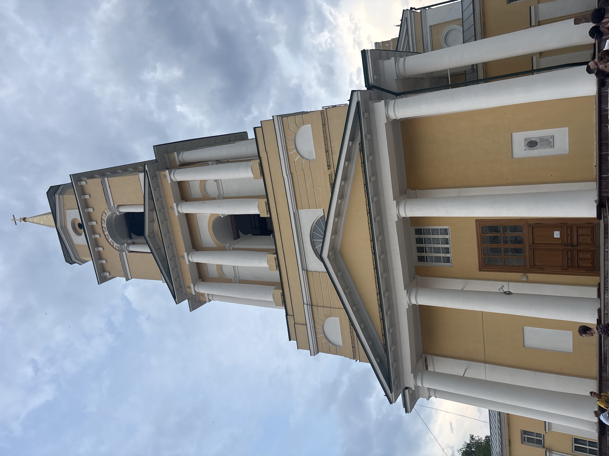 A low-angle, rotated view looking upwards captures a yellow and white building, possibly a church or governmental structure, in Perm, Russia. The building has a multi-tiered tower with columns, arched openings, and a cross atop the highest point of the tower. The main building features large, white, cylindrical columns supporting a roof structure. There are two doors, window panes and a sign. The sky is filled with grey and white clouds. There are a few people visible at the base of the building.