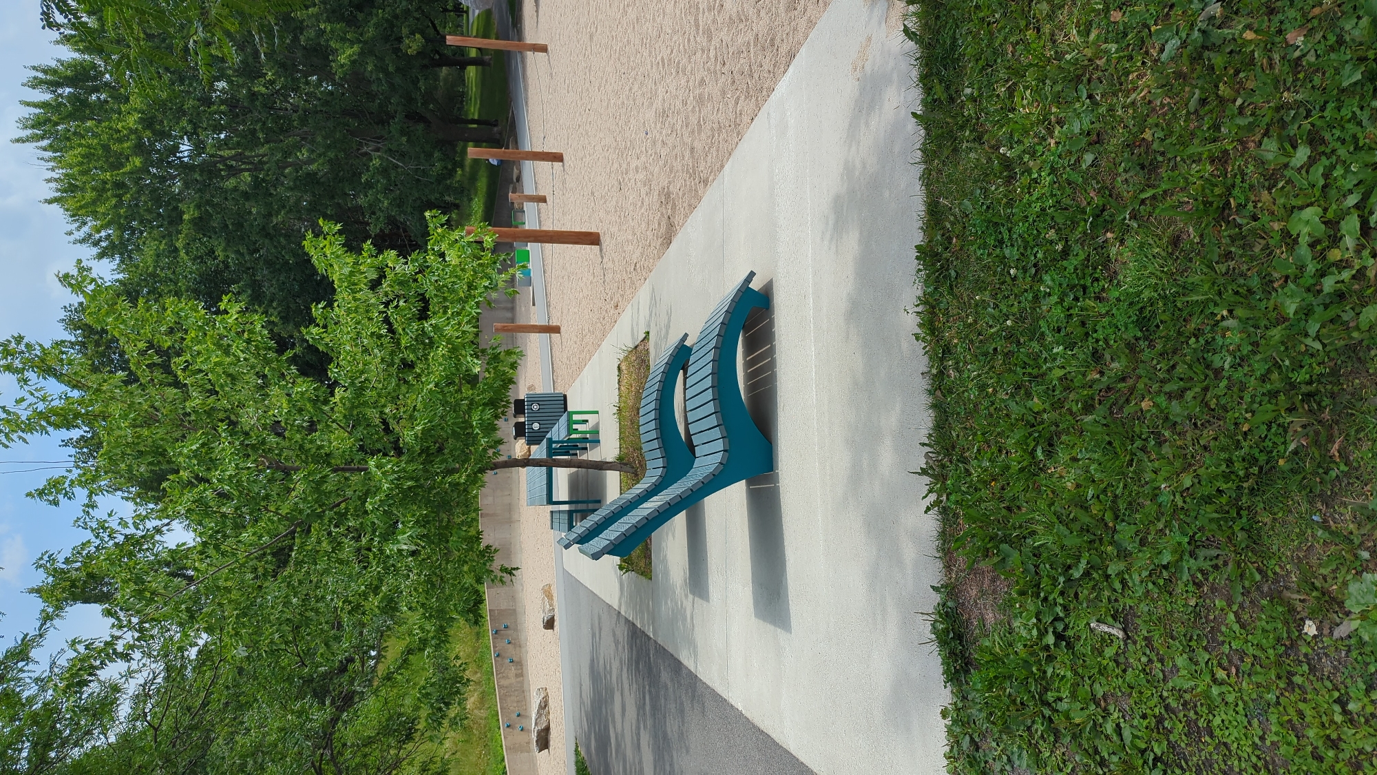 This image depicts an outdoor park scene. In the foreground, a concrete pathway runs horizontally across the frame, bordered by grass on the lower portion. Two teal-colored, curved lounge chairs are positioned on the pathway. Behind the chairs, a teal picnic table with benches is situated under the shade of a leafy green tree.
Further in the background, there is a sandy area, indicative of a playground or recreational space. Several wooden posts are scattered across this sandy section. Beyond the sandy area, a dense line of mature green trees forms a natural backdrop. The sky above is partly cloudy, with a clear blue sky visible.
The scene appears to be a public park designed for recreation and relaxation, likely within a park in Montreal, Canada, given the presence of these amenities and the characteristic greenery. There are no people directly visible interacting with the objects.