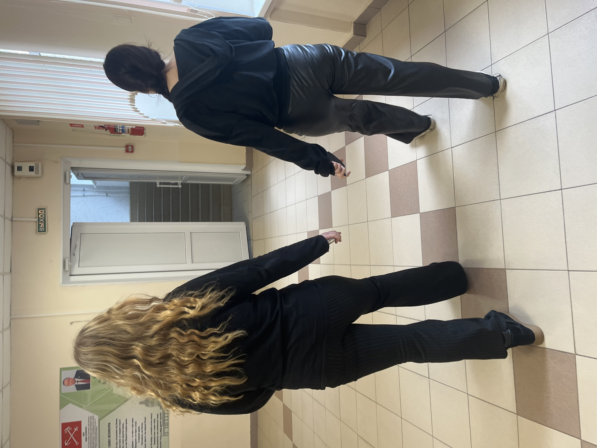 Two women stand in a hallway with a tiled floor and light-colored walls, likely inside a building in Saint Petersburg, Russia.
One woman, with dark hair, wears a black hooded sweatshirt and black pants. She is positioned closer to a doorway with an "EXIT" sign.
The other woman, with blonde, wavy hair, wears a black top and black pants. Behind her is a bulletin board with a picture of a man and some text, possibly related to the city of Saint Petersburg.
The hallway has white doors and a fire extinguisher visible near the ceiling.