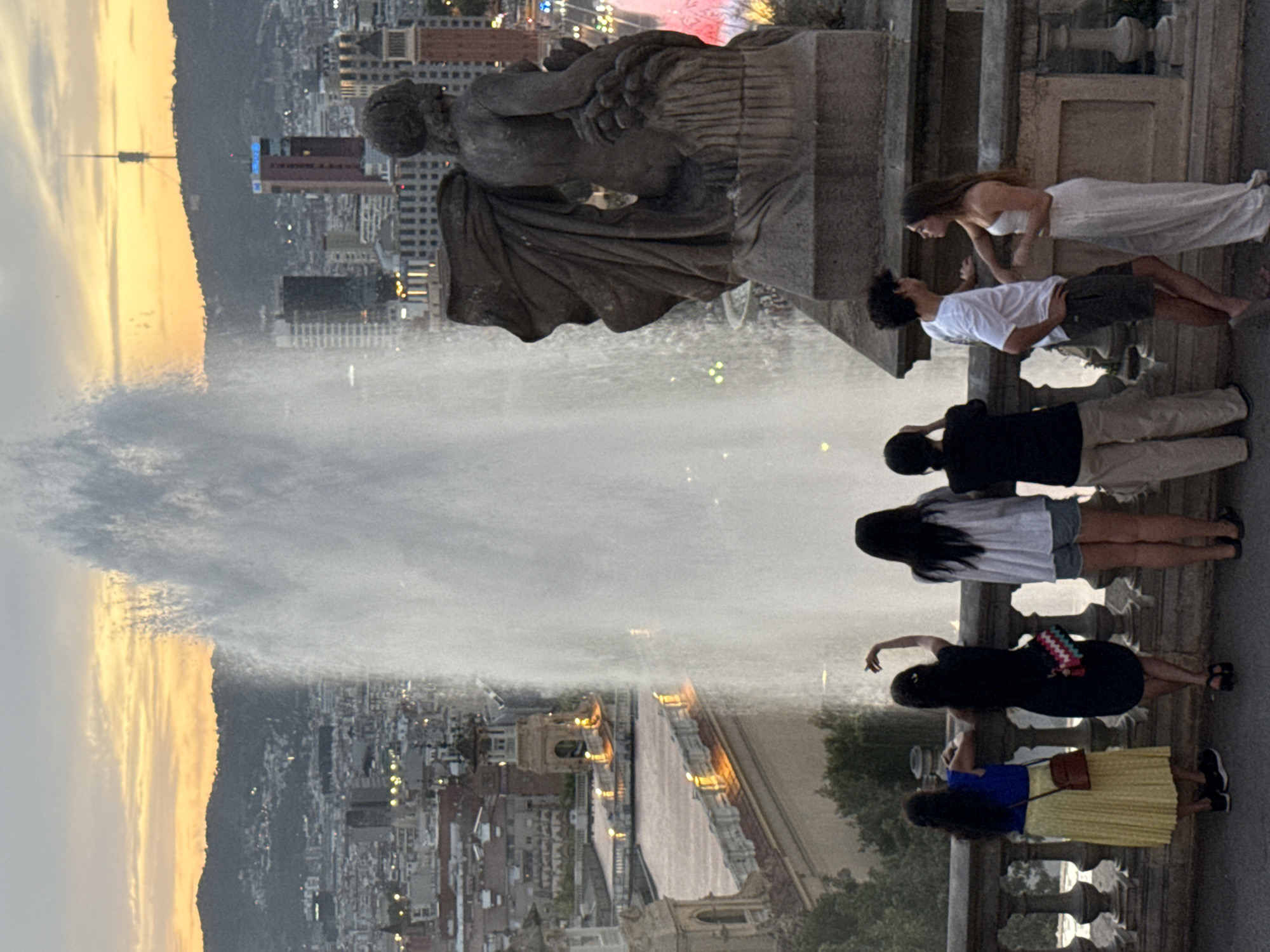 This is a medium shot, eye-level photograph of a group of people standing on a balcony overlooking a city at dusk. The scene is likely taken from Montjuïc, overlooking the city of Barcelona, Spain, with the Magic Fountain of Montjuïc prominently featured as a large water plume in the center of the frame.
On the balcony, from left to right, are six individuals. The first person, a woman, is wearing a bright blue sleeveless top and a long yellow pleated skirt, with a brown purse slung across her body. Next to her, a woman in a dark dress has a colorful bag and her hand raised in a gesture. To her right, a woman in a white t-shirt and shorts stands with her back to the camera, while a fourth person, a man in a white t-shirt and dark shorts, leans forward. He is looking at a woman in a white outfit standing to his right. The sixth person, in a black shirt and tan pants, stands to the far right, partially obscured by the man in white.
In the background, the city of Barcelona stretches out below, with buildings of various heights and architectural styles visible. The sky is a gradient of orange and yellow, indicating sunset or twilight, with a prominent antenna tower on a distant hill. A large stone statue, partially visible on the right side of the frame, adds to the architectural richness of the scene. The lighting suggests artificial illumination within the city and along the balcony itself.