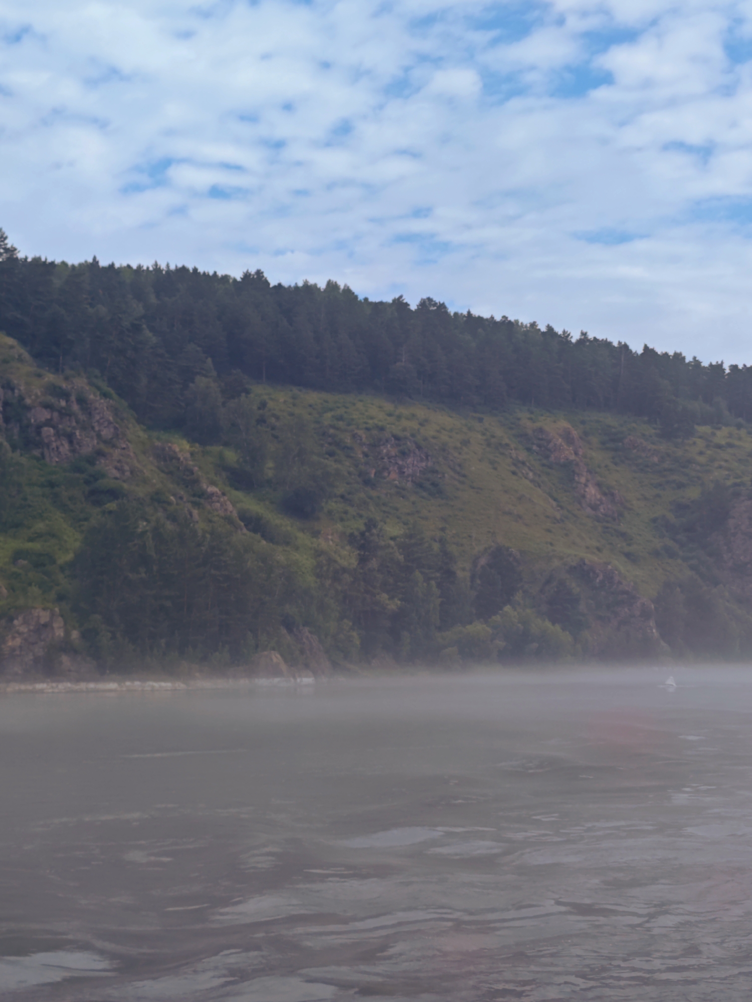 A wide shot captures a misty river landscape against a backdrop of a forested hill. The sky above is a soft blue, dotted with scattered white clouds. The hill, covered in a dense, dark green forest on its upper portion and lighter green vegetation and rocky outcrops on its lower slopes, dominates the upper half of the image. A thick layer of mist hovers over the river in the foreground, obscuring the water's surface in places and creating a soft, hazy atmosphere. A small, white boat is visible in the distance on the river, partially obscured by the mist. The overall scene depicts a serene, natural environment.