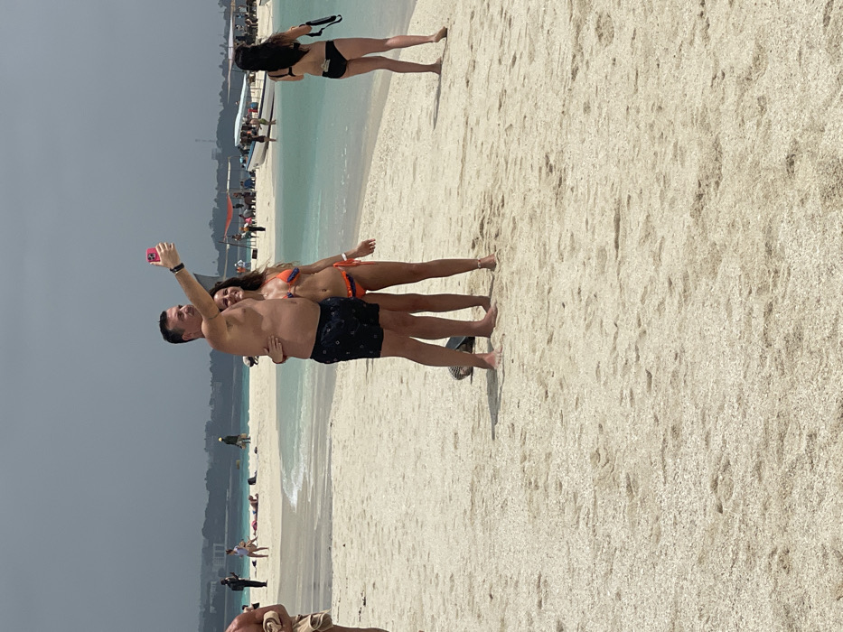 The image depicts a crowded beach scene, likely a sandbank near Stone Town, Zanzibar, Tanzania, characterized by light-colored sand and clear turquoise water under a partially overcast sky. In the foreground-center, a Caucasian man, shirtless and wearing dark swim trunks, holds a pink smartphone at arm's length to capture a selfie. He smiles towards the camera, with his left arm around the shoulder of a Caucasian woman. The woman, wearing an orange and blue two-piece bikini, smiles and looks towards the smartphone, her right arm around the man's waist. Partially obscured dark sandals are visible on the sand near their feet. To the mid-right, a woman in a black two-piece bikini walks away from the viewer, holding a black object in her left hand, with a white rectangular item tucked into the back of her bikini bottom. To the mid-left, a shirtless man's torso and arm are visible, holding a light-colored towel. The mid-ground and background feature shallow, calm water with multiple boats, including a larger white vessel with several people aboard. Numerous other individuals are scattered across the sand and in the water, some lounging, some standing, some obscured by distance. A distant landmass, likely the main island of Zanzibar, is visible on the horizon, showing some structures and vegetation beneath a hazy sky.