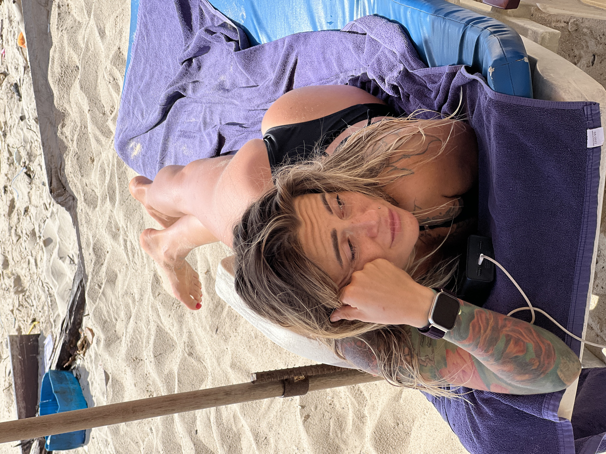 A woman lies on her stomach on a blue lounge chair covered with a purple towel on a sandy beach. She is wearing black bikini bottoms. Her face is turned to the left, with her left hand supporting her head. She has blonde hair that is wet and falls around her face and shoulders. She has tattoos on her left arm and shoulder. A smartwatch is visible on her left wrist. A black portable charger is next to her left arm, connected to a white cable. In the background, there is sand, a wooden pole from an umbrella, and a blue plastic container.