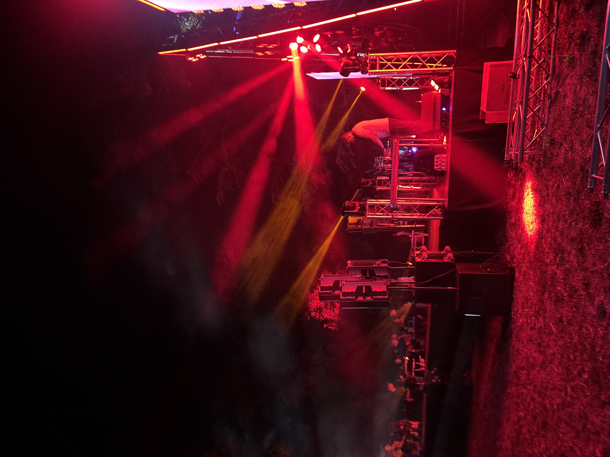 The image shows an outdoor nighttime event with a DJ performing on a stage. The DJ, a person with short hair wearing a light-colored long-sleeved shirt and dark pants, is standing behind a table with audio equipment and operating a mixer. They are illuminated by bright red and yellow stage lights that cast dramatic beams across the scene and into the dark sky. Several people are seated in the background, appearing to be an audience, though their details are obscured by darkness and stage lighting. The stage itself is constructed with metal trussing, and speakers are visible on the left. In the foreground, the ground is covered in grass, also illuminated by red light. In the lower right of the image, metal scaffolding is laid out on the ground, possibly for further stage construction or as part of the event setup. The overall atmosphere is dark and energetic, dominated by the vibrant lighting and the performance.