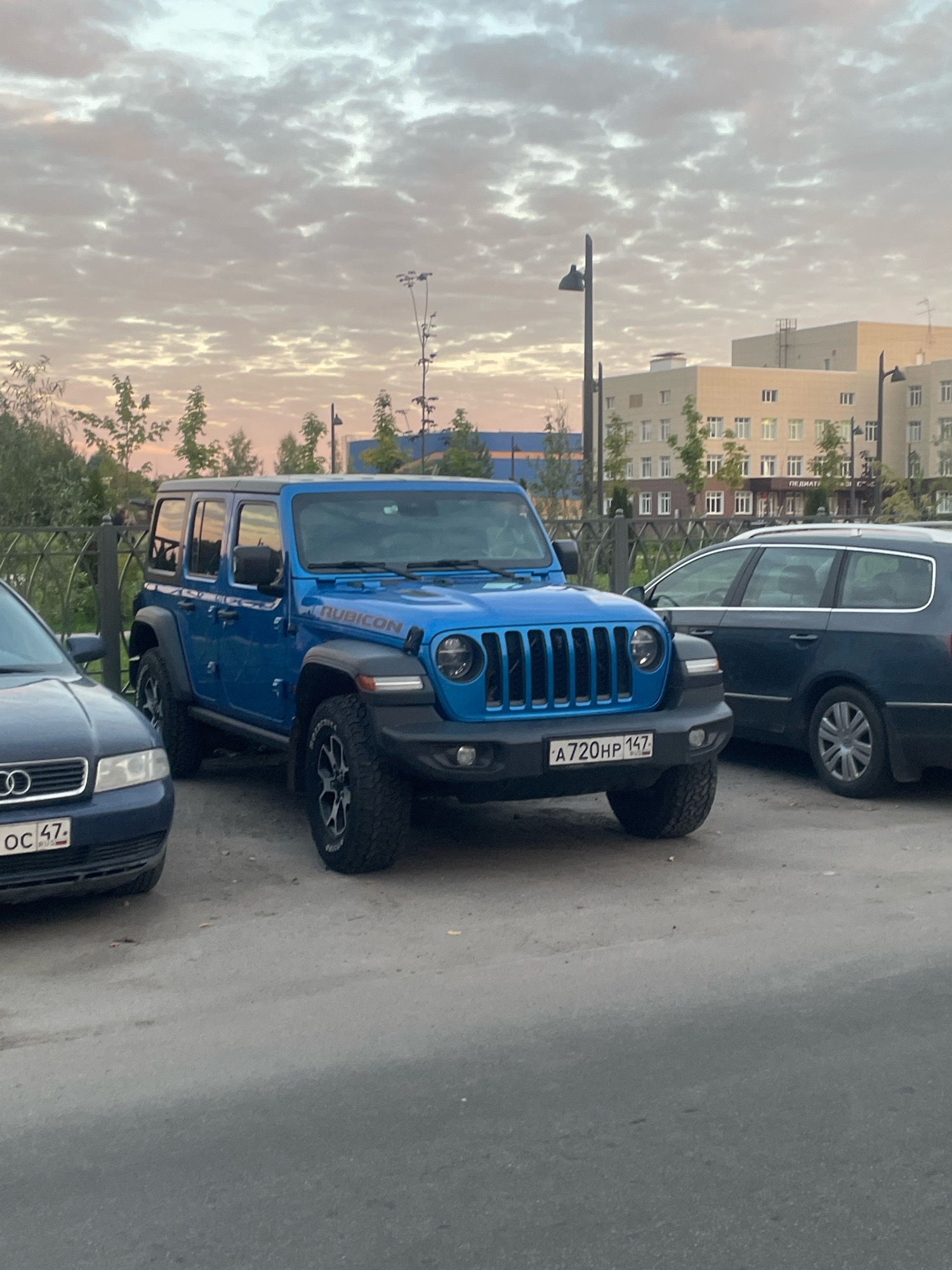 An outdoor, eye-level, medium shot captures a bright blue four-door Jeep Wrangler Rubicon parked on a paved surface, likely a street or parking lot, in Gatchina, Russia. The Jeep is the central subject, positioned at an angle facing slightly left of the camera. It is adorned with the "RUBICON" decal on its side. The Jeep's license plate is visible and reads "A720HP 147". To the left of the Jeep, a dark blue Audi A4 sedan is partially visible, with its front bumper and license plate showing "OC 47". To the right of the Jeep, a dark colored station wagon is parked, with its rear section visible. In the background, a light-colored, multi-story building with many windows stands prominently. Further in the background, there are trees and a fence. The sky above is filled with a patterned cloud formation, suggesting a twilight or early morning setting, with soft, diffused light. The overall scene depicts a common urban or suburban parking scenario under a clouded sky.