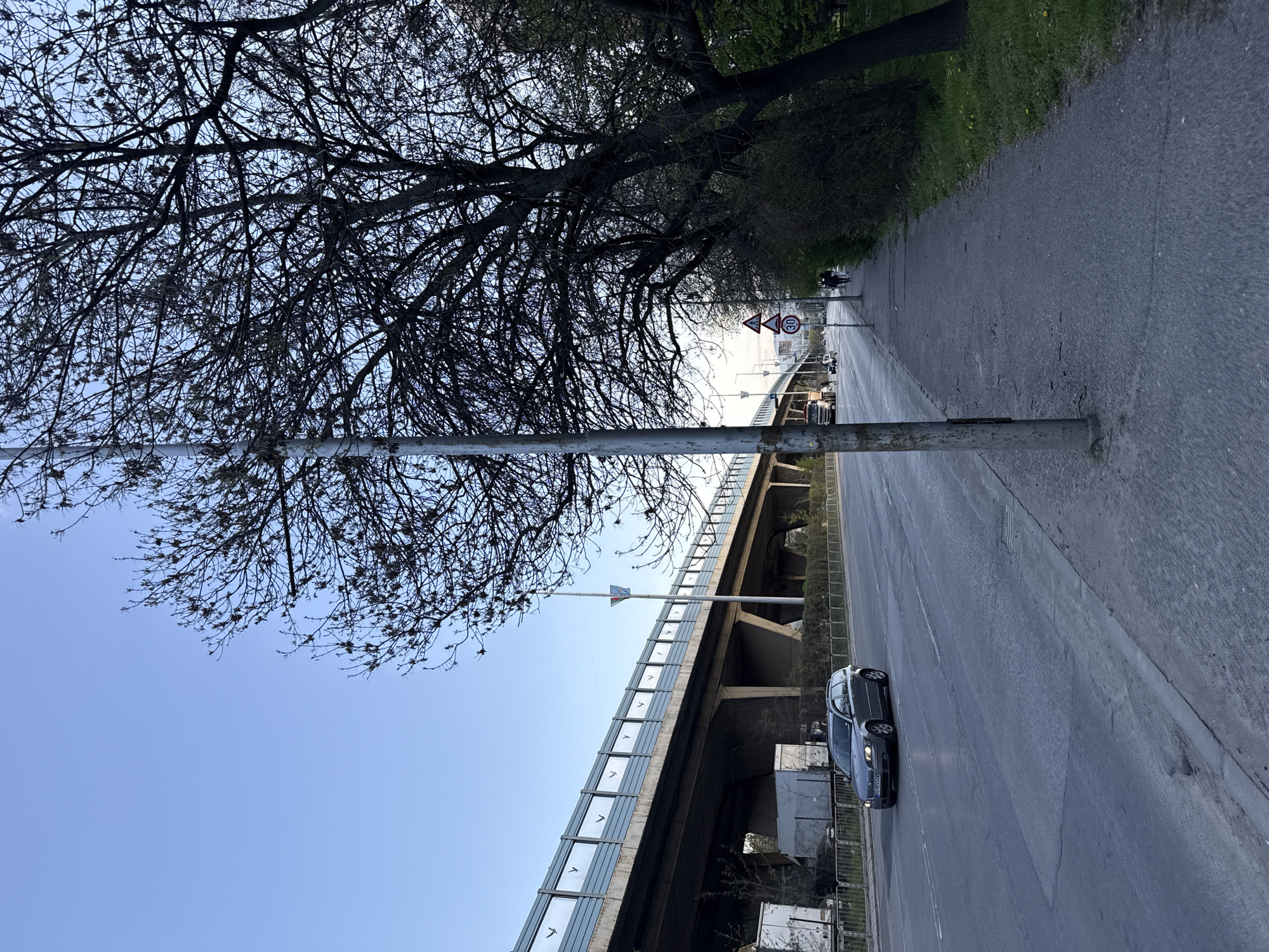 The image shows a street scene in Sofia, Bulgaria, with a road, a sidewalk, a bridge, and a tree. A dark gray car is driving on the road. A metal pole stands between the road and the sidewalk. A highway overpass spans the street. A few people are walking on the sidewalk in the distance. The sky is a light blue.