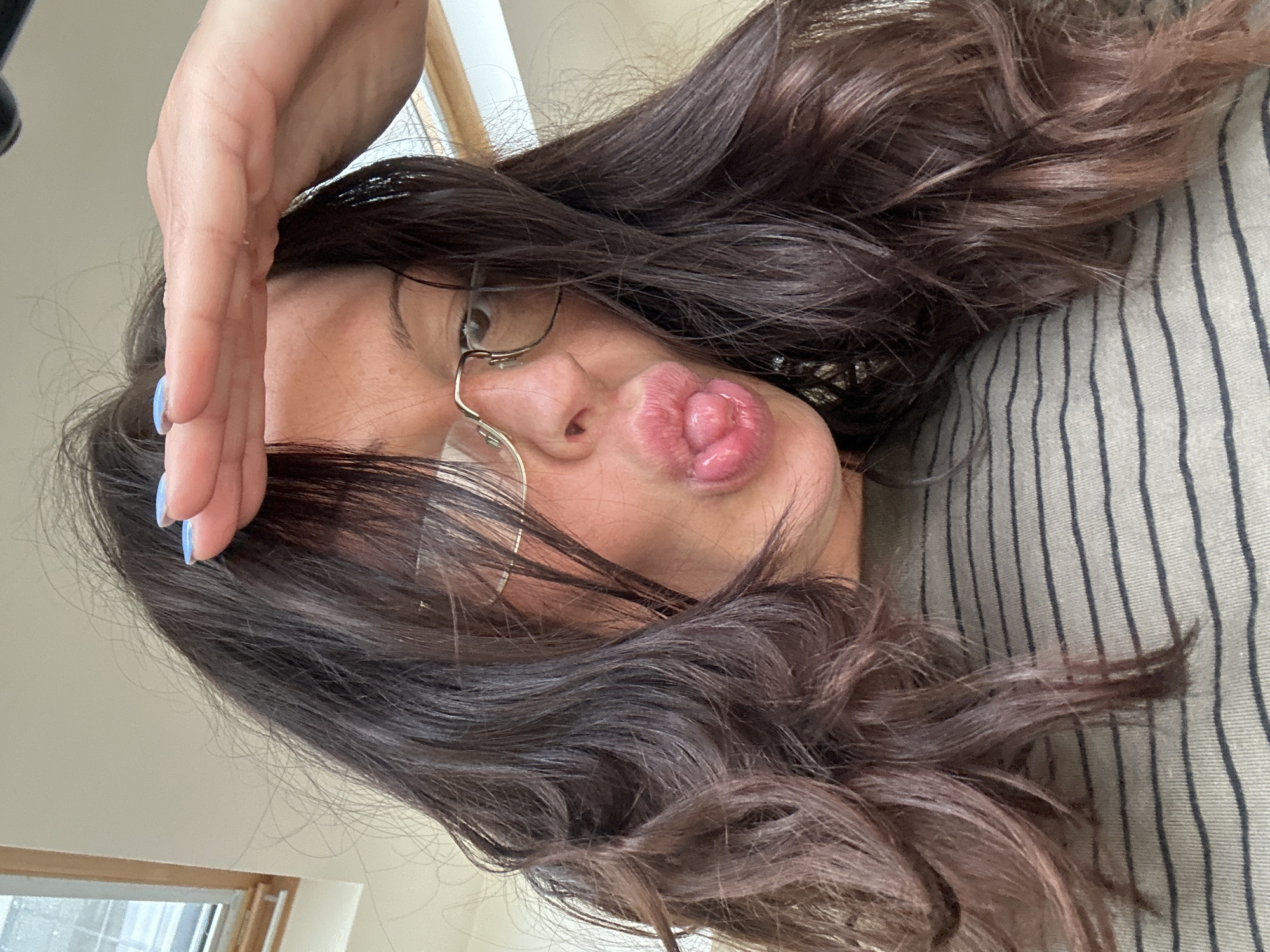 A woman with long, dark, wavy hair and glasses is lying down, appearing to be resting on a striped fabric surface. She has her hand near her forehead and is puckering her lips. A window is visible in the background, suggesting an indoor setting. The location in the city of Tunbridge Wells, United Kingdom is not determinable from the image.