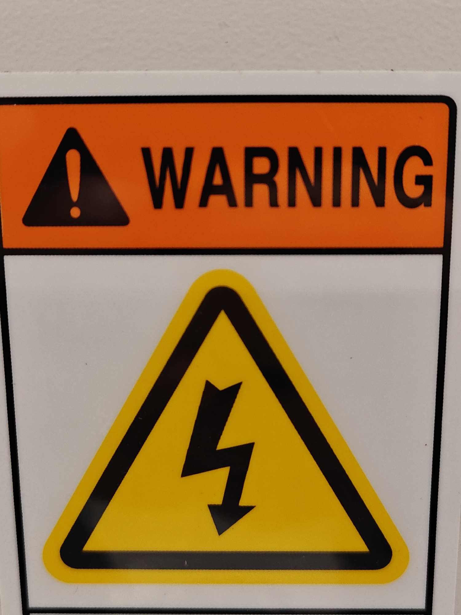 The image shows a close-up of a warning sign. The sign has two distinct sections. The top section is an orange rectangle with a black border. Inside the orange rectangle, there is a black equilateral triangle with an exclamation mark inside. To the right of the triangle, the word "WARNING" is written in large, black, sans-serif capital letters. The bottom section of the sign is a white rectangle with a black border. Inside this white rectangle, there is a large yellow equilateral triangle with a black border. Inside the yellow triangle, there is a black lightning bolt symbol pointing downwards. The sign appears to be mounted on a white surface, possibly a wall or an electrical enclosure. The lighting suggests an indoor environment. There are no people or any other identifiable objects or actions in the image.