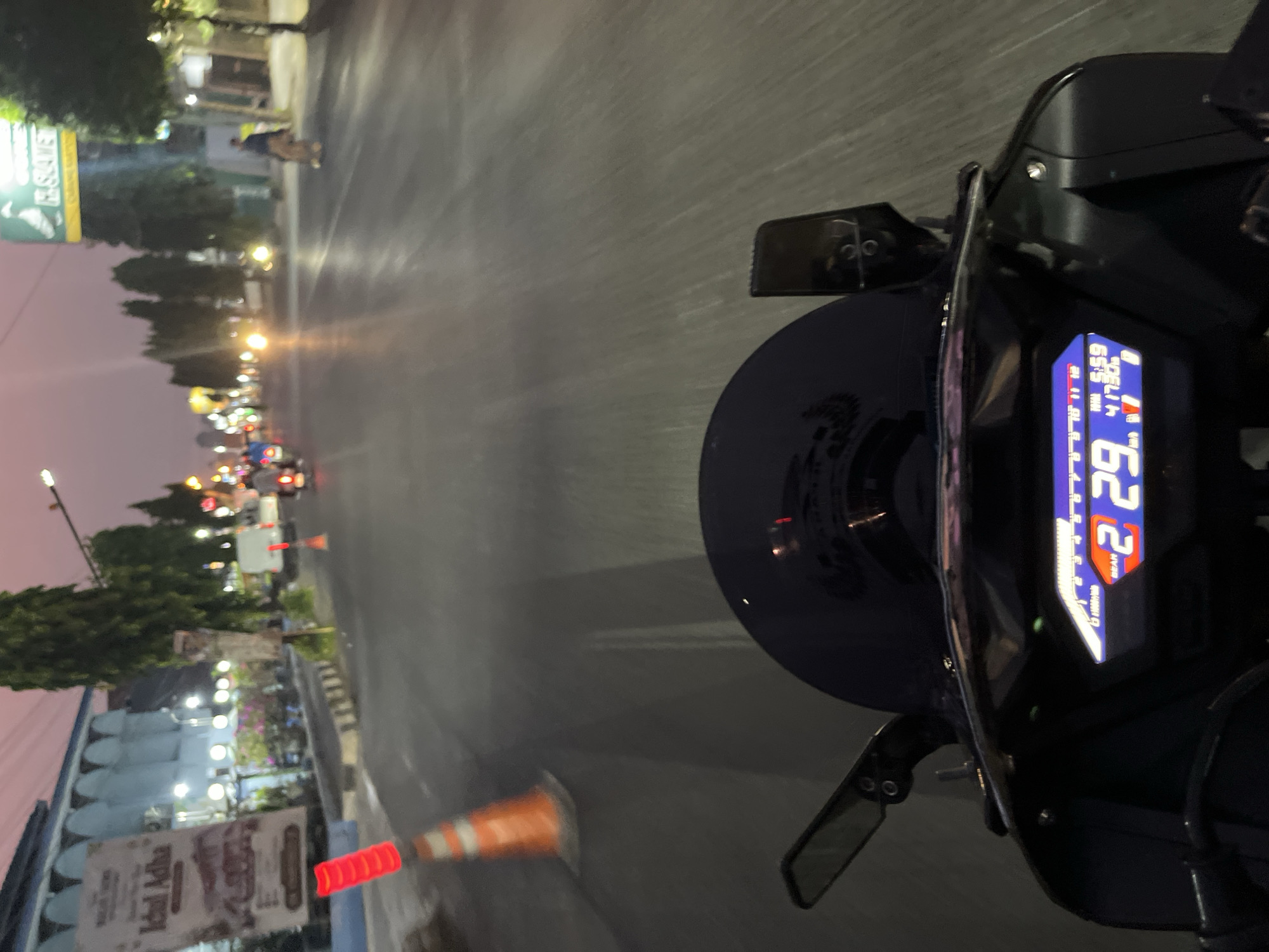 The image depicts a point-of-view shot from a motorcycle traveling on a road in Kalinyamatan, Indonesia, during twilight. The motorcycle's digital dashboard is visible in the foreground, showing a gear indicator of '2', a speed reading of '29' km/h, and the time as '5:59' with an odometer reading of '41730' km. The road ahead is paved and appears wet, reflecting the streetlights and vehicle lights from the surroundings. Several traffic cones with reflective red tops are placed along the side of the road, indicating construction or a hazard. On the left side of the road, a building with a light blue facade and arched windows is visible, with posters displayed on its exterior. Trees line the street, and various vehicles, including motorcycles and a white van, are present in the distance. A pedestrian is also visible on the right side of the road. The sky displays a soft pink and orange hue, characteristic of sunset or sunrise. The overall scene suggests a typical urban environment at dusk in Indonesia.