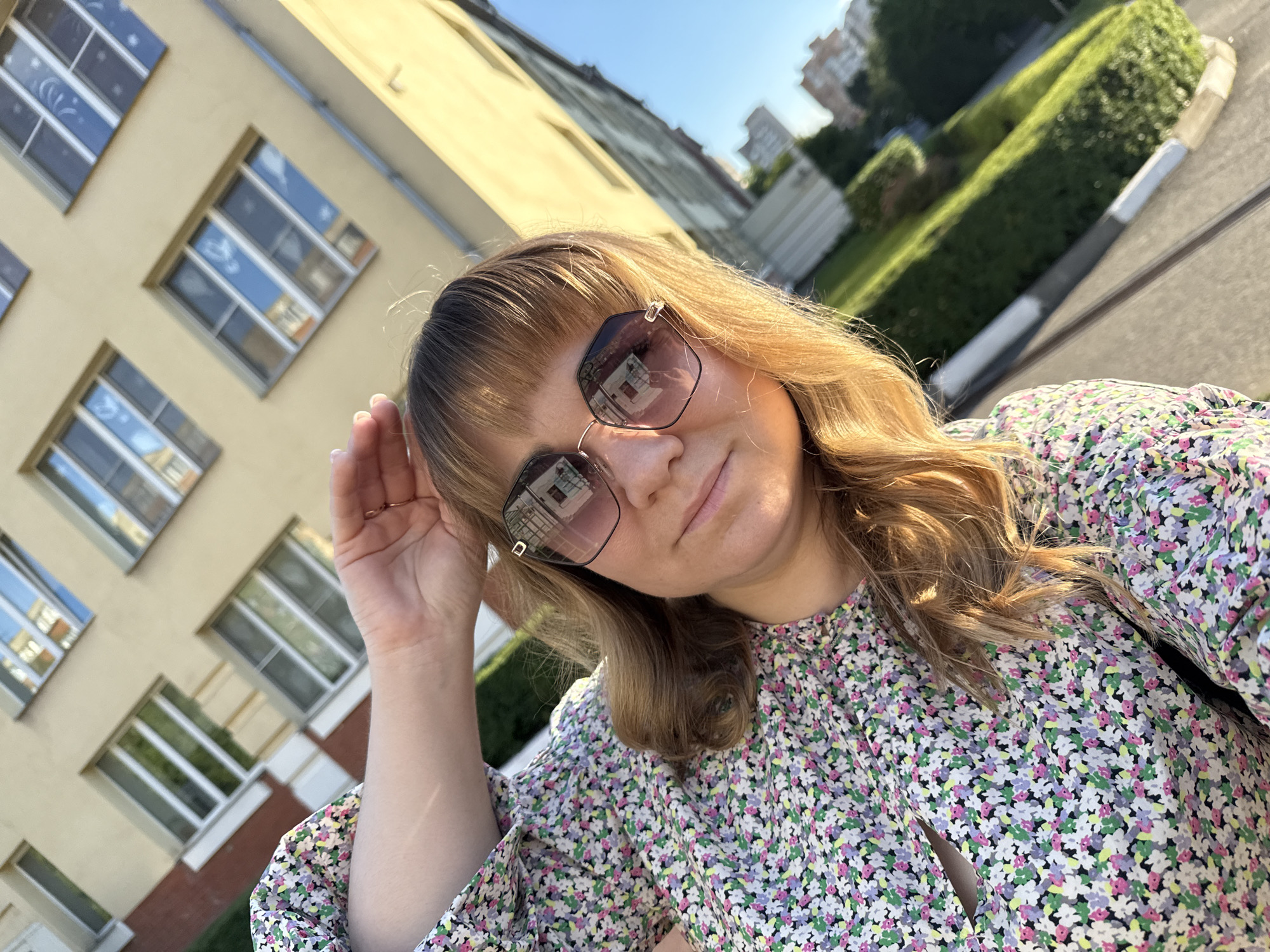 In this selfie, a woman with blonde hair and bangs is wearing sunglasses and a floral print dress. She is holding her hand up to her hair. The background features a light-colored building with several windows and a well-maintained green lawn. The sky is clear and blue, suggesting a sunny day. Buildings and trees are visible in the distance. The scene appears to be outdoors, likely in a residential or school area. The lighting indicates it is daytime, possibly late afternoon due to the warm sunlight. The overall mood seems relaxed and casual. There is no visible text in the image.