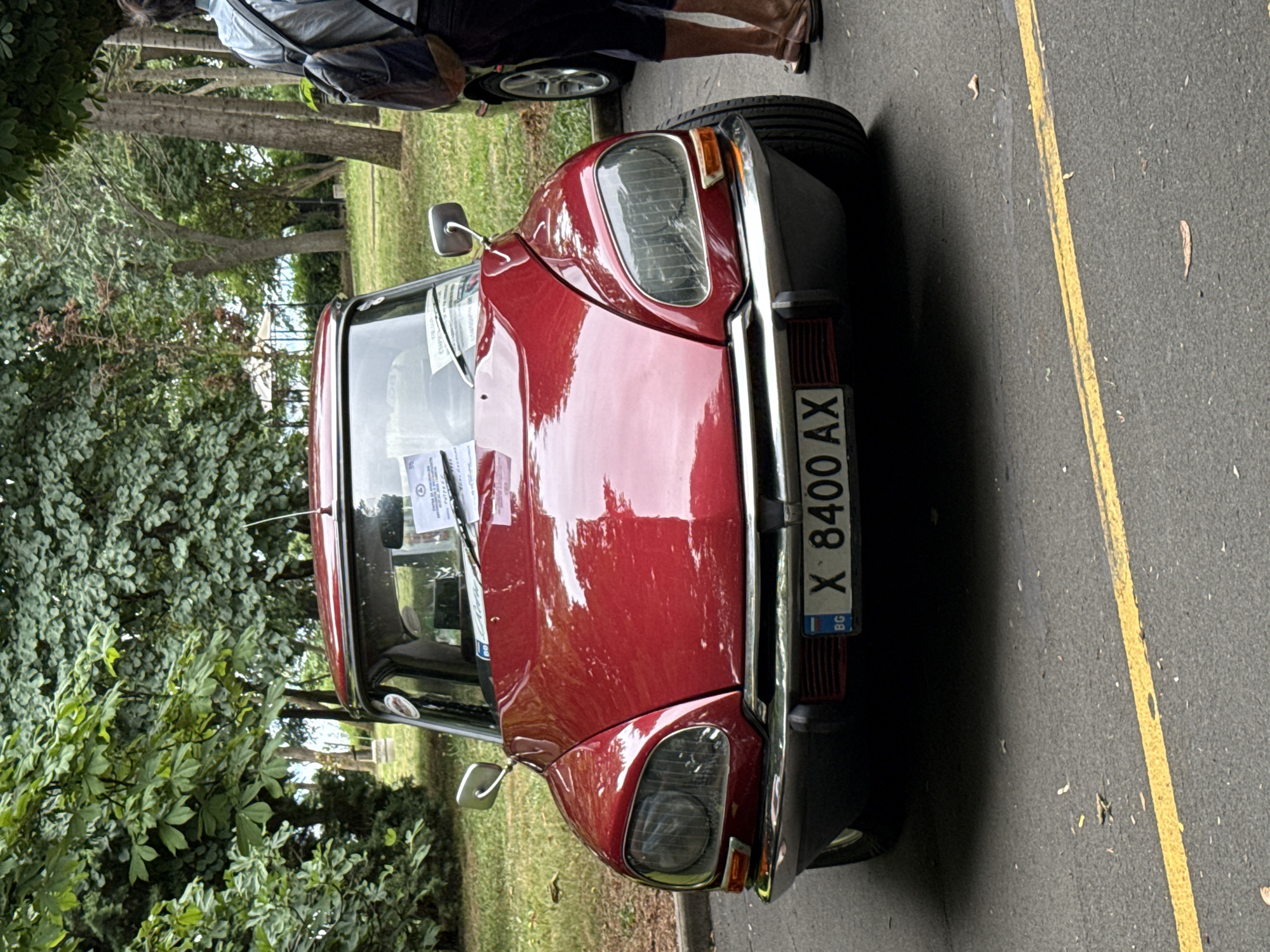 The image shows the front view of a red vintage car, likely a Citroën DS, parked on an asphalt road with a yellow line. The car has a chrome bumper and distinctive large headlights. A Bulgarian license plate with the characters "X 8400 AX" is visible. In the background, lush green trees and foliage create a park-like setting. To the right of the car, the legs of a person wearing dark shorts and sandals are visible, along with a backpack slung over their shoulder. The overall scene suggests an outdoor event or car show. Burgas, Bulgaria is known for its parks and outdoor events, making this setting plausible.