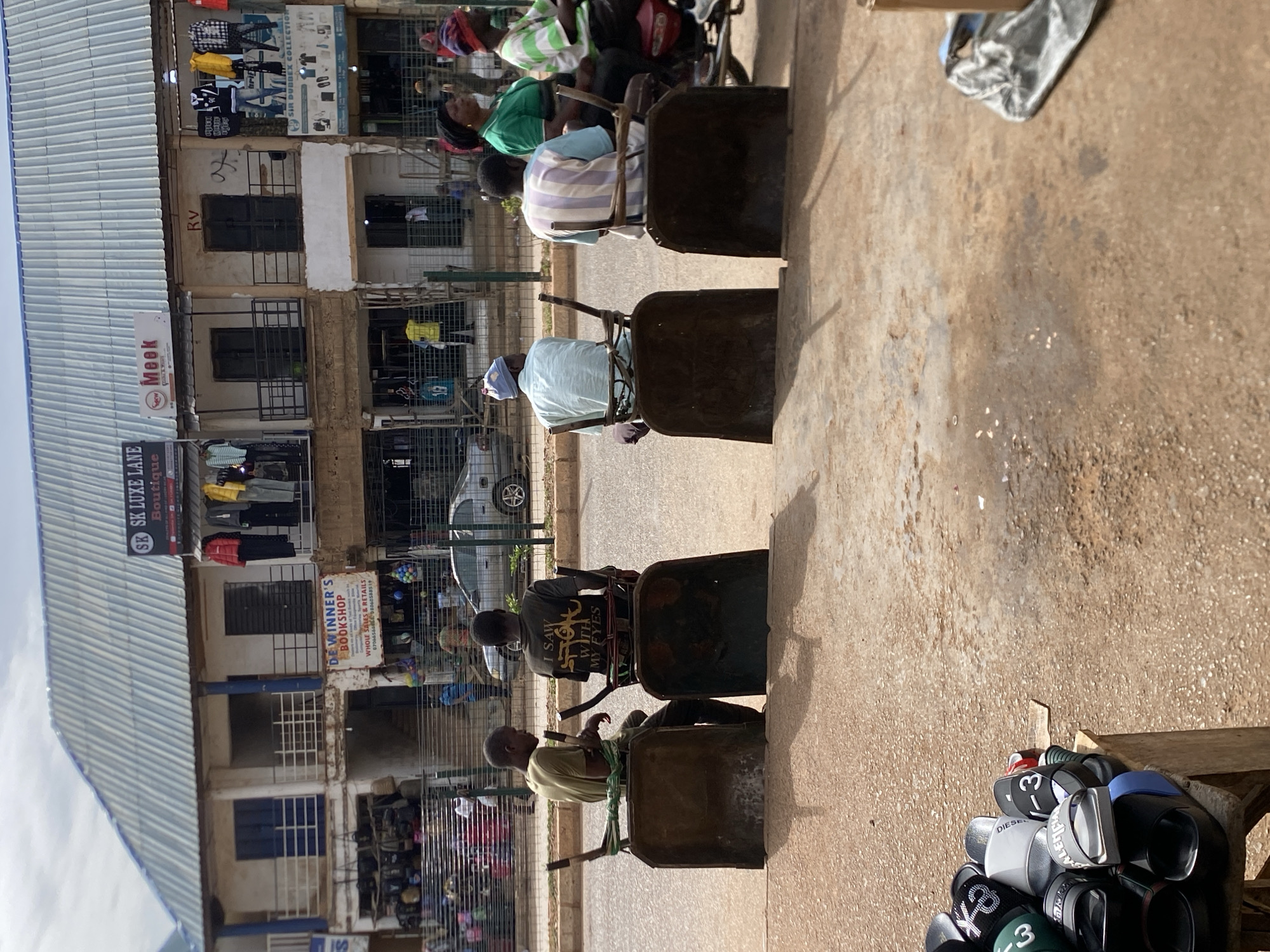 The image depicts a street scene in Bukuru, Nigeria. Several individuals are seated in modified wheelbarrows that function as chairs, lined up along the side of the road. A multi-story building with shops is visible in the background. The buildings showcase signs like "SK LUXE LANE Boutique," "DE WINNER'S BOOKSHOP," "Meek," "Rv," and "SIR DUBBEX COLLECTION." Clothes hang on display in some of the shops. A car is parked on the roadside. Additionally, a pile of footwear, including sandals and shoes, sits on a small wooden table on the edge of the sidewalk. The road is made of a packed brown stone mix.