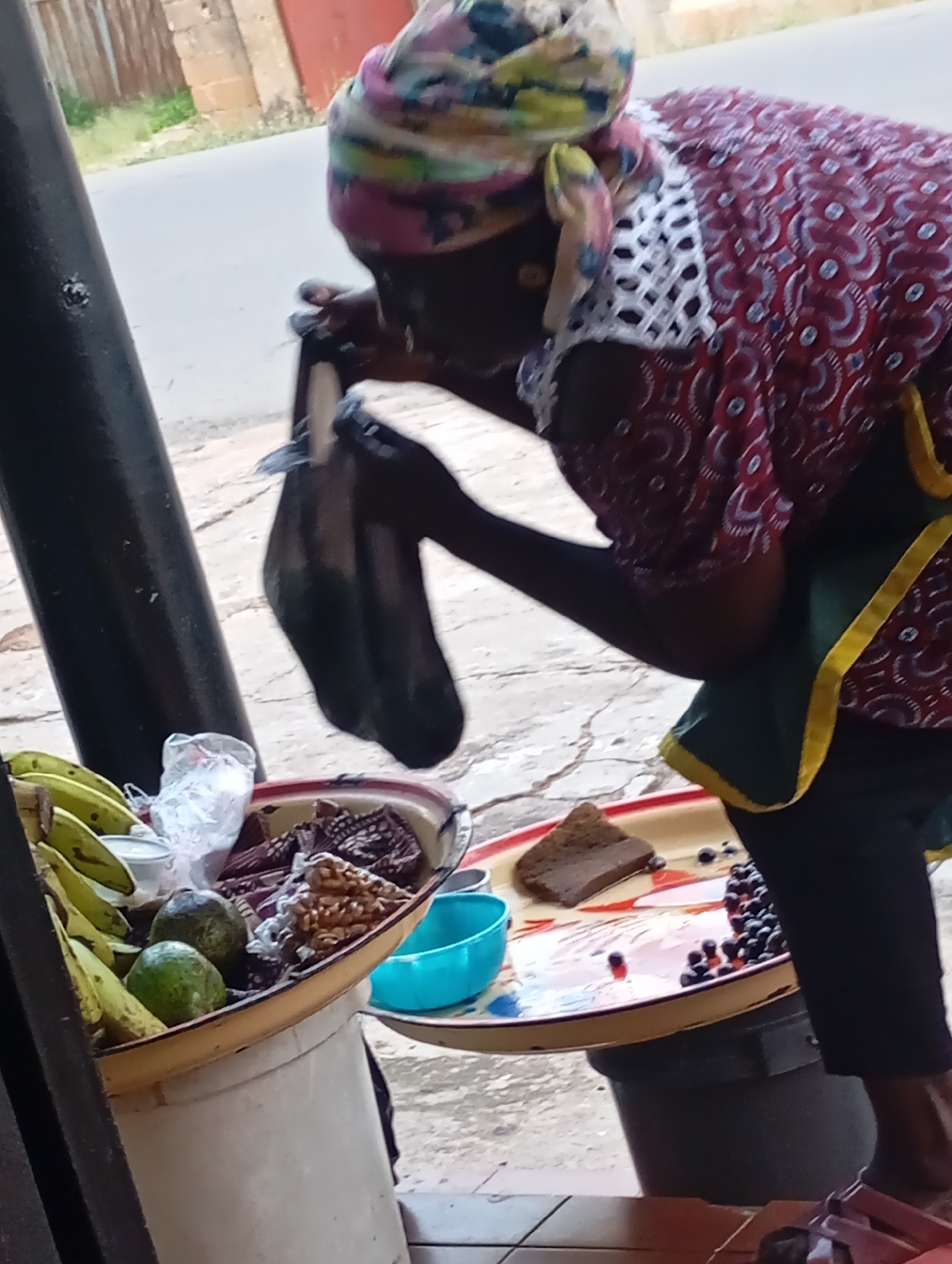 The image shows a person, likely a woman, in a vibrant red patterned garment and a colorful headscarf, bending over a selection of goods displayed for sale. She is wearing a green apron with yellow trim. She is holding a dark plastic bag, appearing to be either retrieving or placing something inside it.
The goods are arranged in various containers. To the left, a large, pale bucket holds a bunch of green bananas, two avocados, and some items wrapped in plastic and patterned fabric. Next to this, a shallow, round tray with a red rim holds a slice of dark bread, a small blue bowl, and a pile of small, dark berries. The tray is placed on top of a dark cylindrical object, possibly a bin or cooler.
The background suggests an outdoor setting, likely a roadside stall or market in Jos, Nigeria. A paved road is visible, along with a brick building and a corrugated metal fence. The ground appears to be a textured, light-colored surface, possibly concrete or pavement, with some cracks and debris. The lighting suggests it is daytime. The woman's posture and the presence of goods for sale indicate she is engaged in a commercial activity, likely selling food items. There is no visible text in the image.