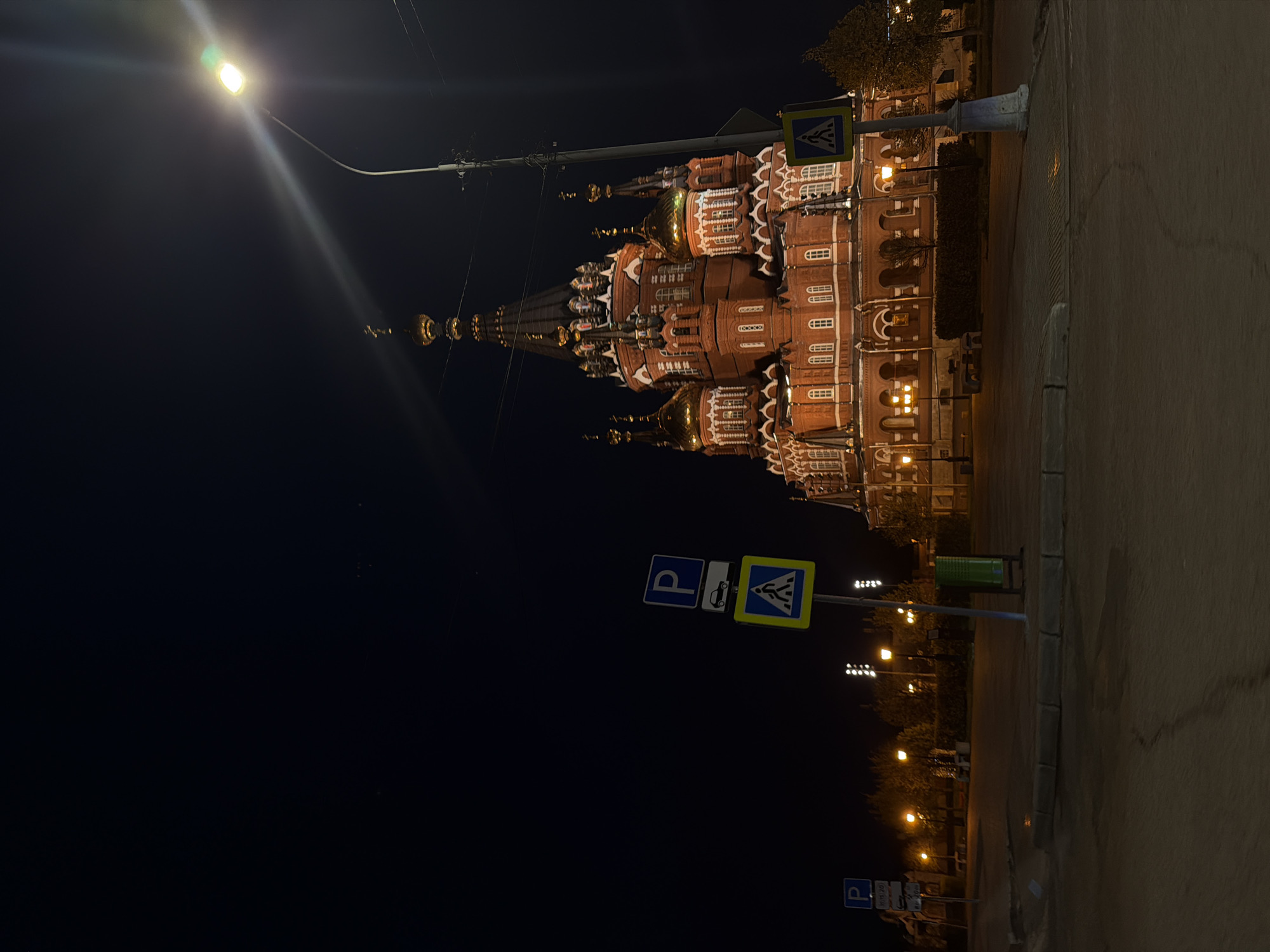 The image shows St. Michael's Cathedral in Izhevsk, Russia, at night. The red-brick cathedral is illuminated with a warm glow, contrasting with the dark sky. A street light shines brightly near the cathedral. A parking sign and a pedestrian crossing sign are visible, along with other street lights in the distance. The image appears to be taken from a low angle, emphasizing the height of the cathedral. The ground is paved and the photo is rotated clockwise by 90 degrees.