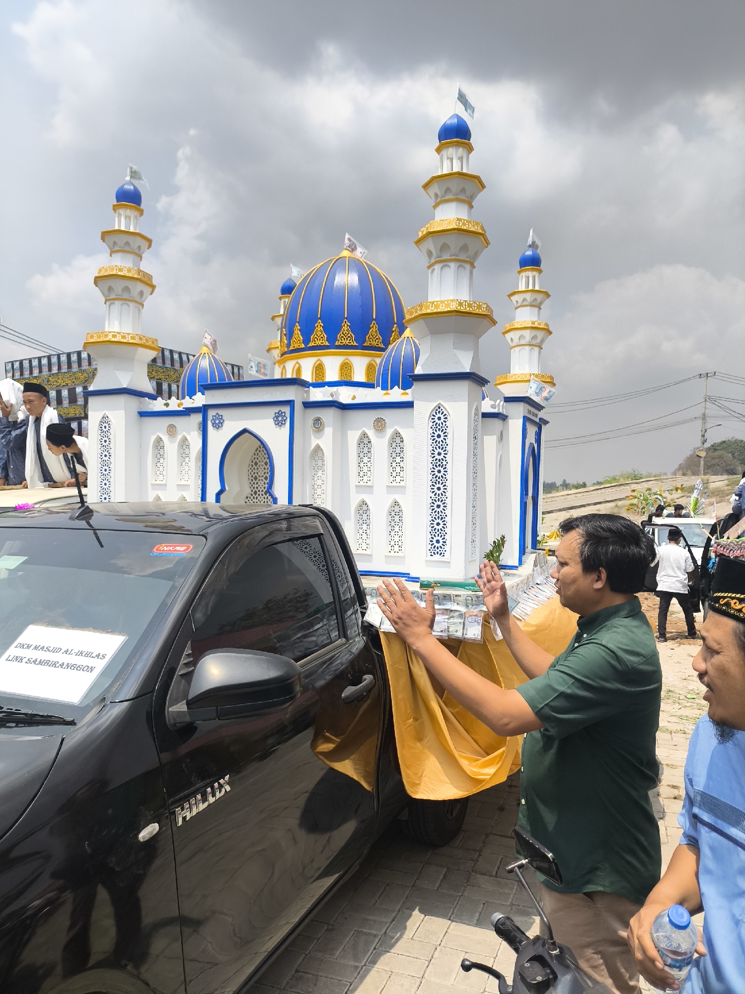 In Cilegon, Indonesia, a miniature mosque replica, decorated with blue domes and white walls, is displayed on a flatbed truck alongside a black Hilux pickup truck. The truck is adorned with a yellow cloth and stacks of money. Two men are present: one in a green shirt is seen placing money onto the yellow cloth, while another man in a blue shirt stands nearby holding a water bottle. Several other individuals are visible in the background near the mosque replica and a white truck. The scene takes place outdoors under a cloudy sky with some vegetation and a road visible in the distance.