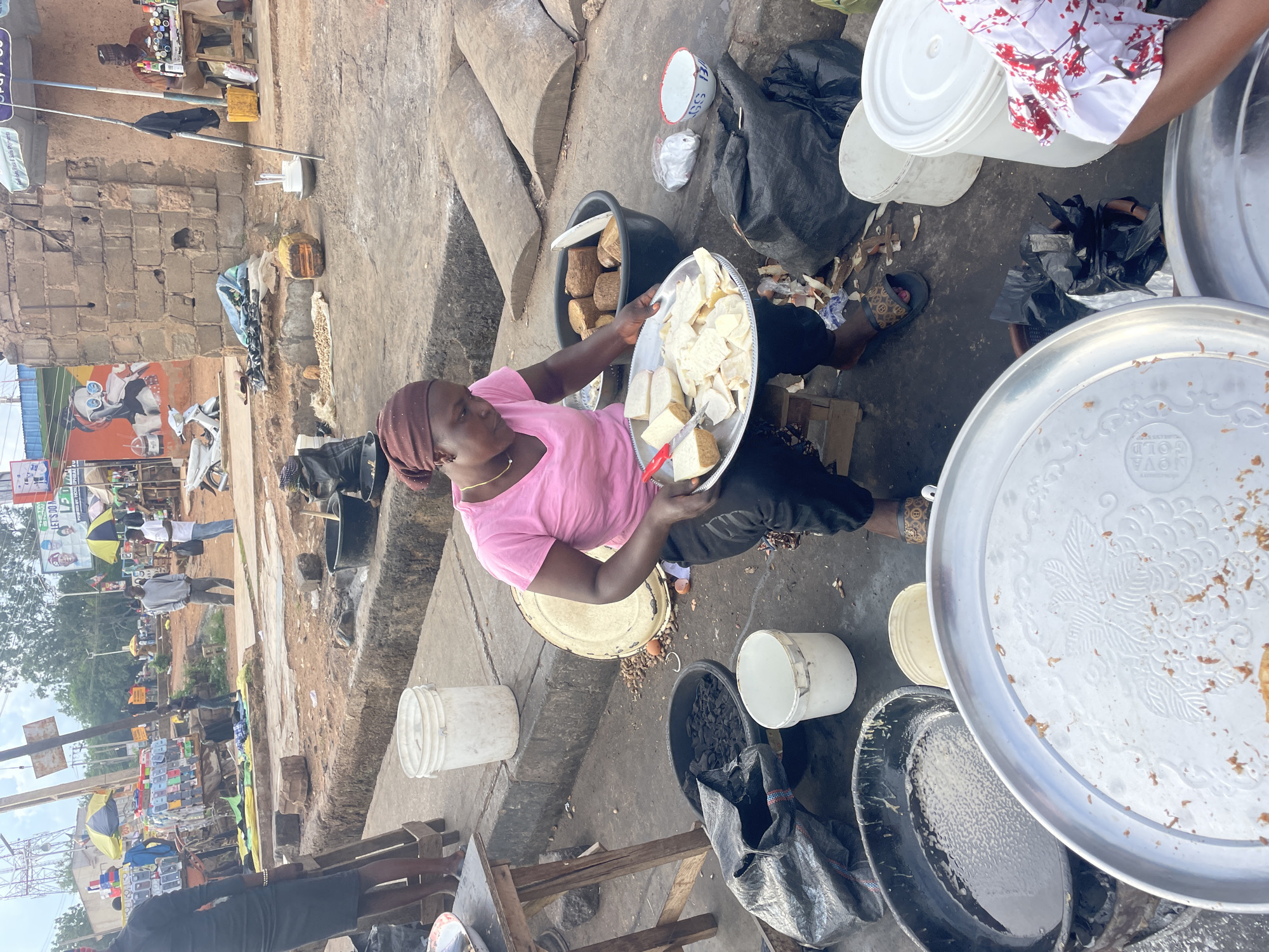 This detailed description pertains to an outdoor market scene in the city of Jos, Nigeria, characterized by informal street vending and pedestrian activity under an overcast sky during daytime.
In the immediate foreground, a dark-skinned woman with a maroon head covering, a pink t-shirt, dark trousers, and a thin gold-colored necklace is seated on a low stool. She holds a large, round silver-colored metal tray with both hands, on which various light-colored, cut food items, possibly tubers like yam or cassava, are arranged. A small red-handled knife rests on the food. Directly in front of her feet is a large black tub containing a frothy, light-colored liquid. To her left (viewer's right) are two stacked, large, round silver-colored trays, the topmost of which is empty and decorated with an embossed floral pattern. To her right (viewer's left), a large black plastic tub holds dark, chunky material, possibly charcoal. Beside it are two white plastic buckets. Behind her, a large round metal lid rests on the ground amidst scattered food scraps, including what appear to be peanuts and eggshells. She wears dark slip-on sandals with a patterned strap.
The midground features a concrete pathway and a shallow, open concrete drainage ditch. Further into the scene, a bustling street environment unfolds. To the far left, a person in a dark dress stands behind a wooden table. Adjacent to them is an informal stall displaying numerous colorful phone cases or similar accessories arranged vertically. Two men stand in the center-right; one wears a grey t-shirt and jeans, the other a white t-shirt and light pants, holding a bag. Several yellow and black striped market umbrellas provide shade over various vendor setups. To the far right, against a rough-hewn brick building, a vendor sits under a blue umbrella, selling small bottles of liquids from a table. A large, brightly colored mural depicting a person with sunglasses and a drink is visible on a building further back on the left.
The background shows additional buildings, some in brick and others with painted surfaces. A billboard in the distance displays text, including "POWERED BY: HON. EPHRA...", possibly indicating local political or business sponsorship. A large metal telecommunications tower rises in the far left background. The ground throughout the scene is a mix of compacted dirt and concrete, showing signs of frequent use and some scattered debris.