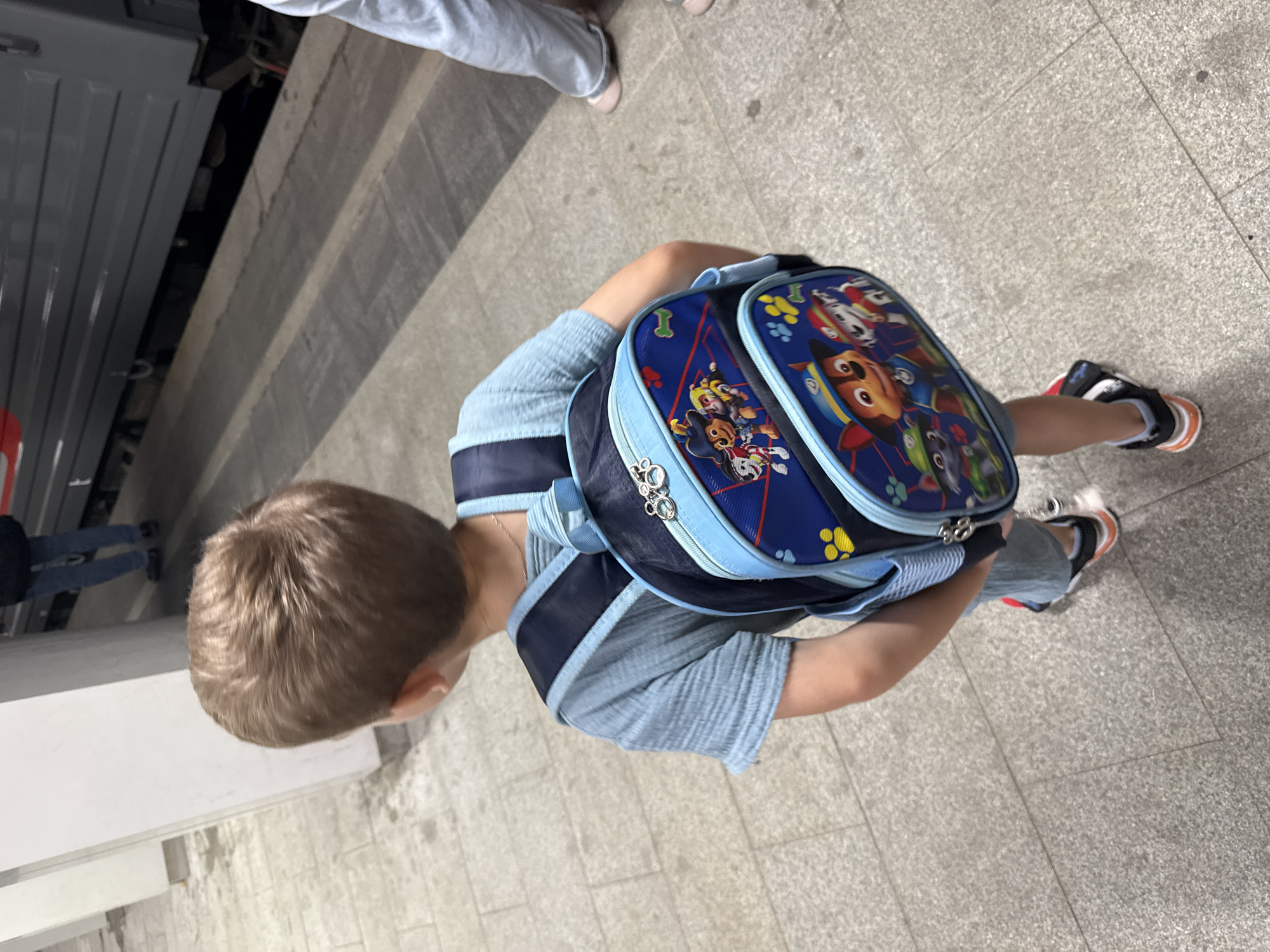 A young boy with light brown hair is seen from behind, wearing a light blue t-shirt and a blue backpack featuring characters from the Paw Patrol franchise. He is standing on a concrete platform, possibly at a train station, with a train visible in the background. His feet are clad in sneakers with orange and black accents. The platform floor is a light gray, speckled concrete. The overall scene suggests a public transport setting. The image was taken in Moscow, Russia.