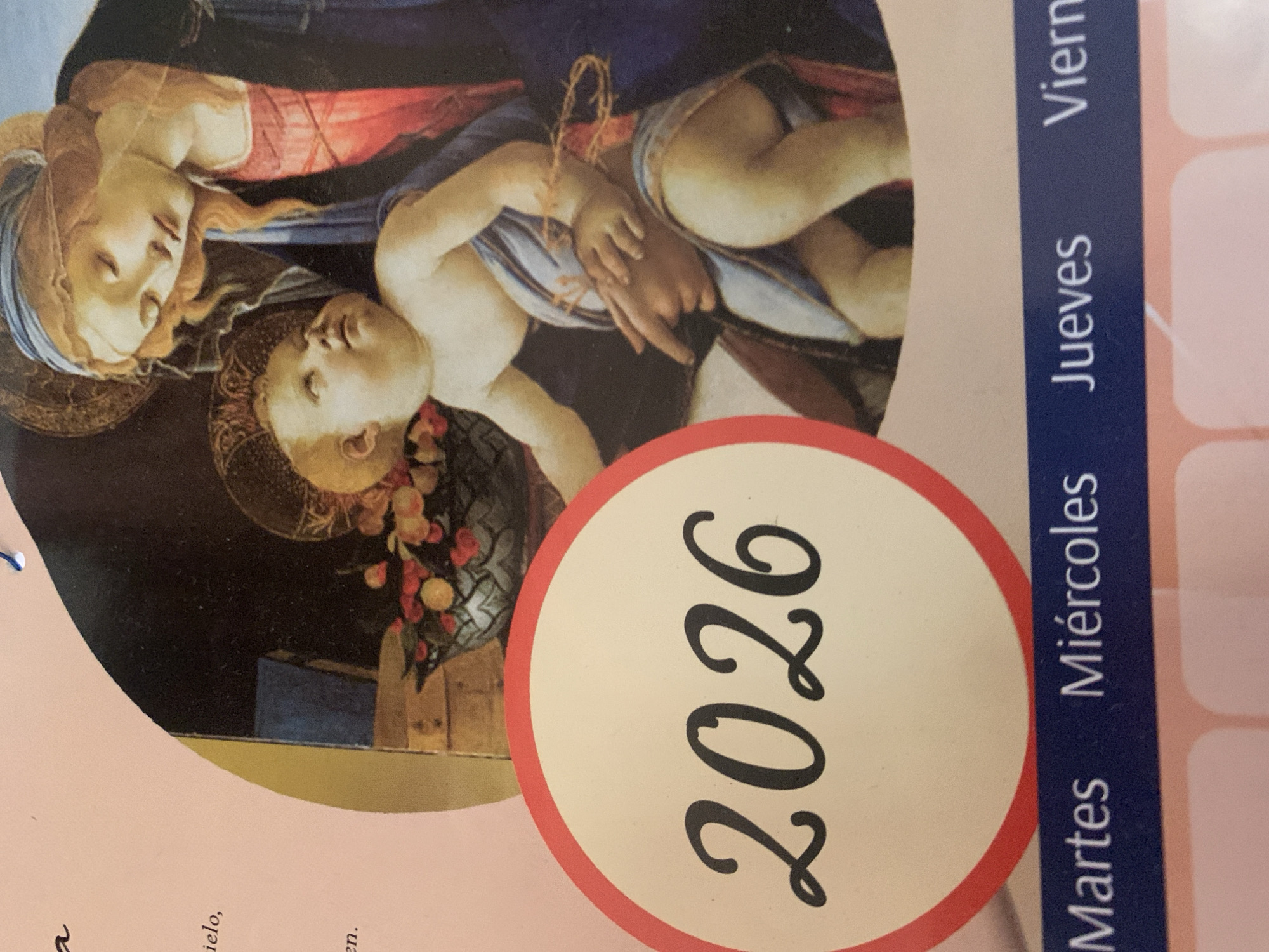 The image displays a tilted, close-up view of a printed item, likely a calendar. The main visual element is a reproduction of a classical painting featuring two figures: a woman and an infant. The woman, possibly the Virgin Mary, has auburn hair, a blue head covering, and a red garment over a blue one, with a faint halo. She gazes downwards with a serene expression. The infant, likely Jesus, is light-skinned, partially covered by a striped cloth, and also has a halo. A small crown of thorns rests on the infant's arm, and a floral or fruit arrangement is visible near their head. The background of the painting is dark.
Prominently featured on the calendar is a large circular label with a red border, containing the year "2026" in elegant black script. To the right of this, a vertical blue band lists days of the week in Spanish: "Martes" (Tuesday), "Miércoles" (Wednesday), and "Jueves" (Thursday), with "Viern" (likely "Viernes" for Friday) partially visible. The surrounding calendar pages are a soft pink color. Small, incomplete Spanish words like "ielo" and "en" can be seen in the bottom left corner. The overall scene suggests a calendar for the year 2026, intended for a Spanish-speaking audience, featuring religious art.