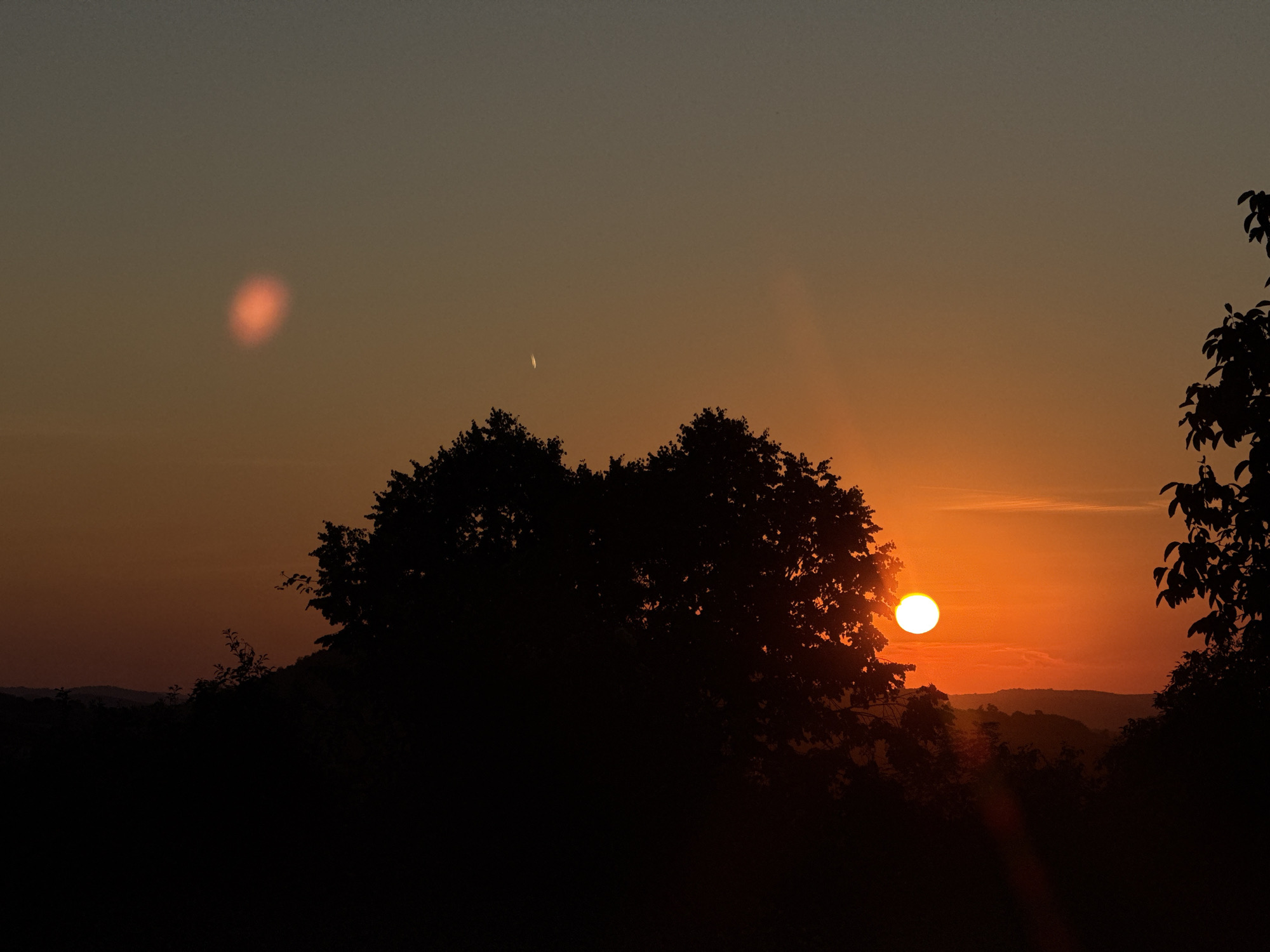 The image captures a sunset over a hilly landscape, silhouetting trees and vegetation against the warm hues of the sky. In the foreground, a large, dark mass of foliage dominates the lower portion of the frame, with the leaves of a tree on the right side of the image extending into the frame. A prominent bright sun, a perfect orb of light, is visible near the center-right, casting a vibrant orange glow. In the mid-ground, rolling hills are faintly discernible under the sunset sky. The sky itself transitions from a deep orange near the horizon to a dusky gray at the top of the image. Notably, there are two distinct light phenomena in the sky: a reddish-orange smudge on the upper left, and a thin, bright streak, possibly a contrail, slightly above and to the left of the sun. The overall impression is of a serene and natural landscape at dusk, with the setting sun being the focal point. The specific location in the city of Krushevo, Bulgaria, is not visually indicated by any landmarks or identifying features within the image itself.