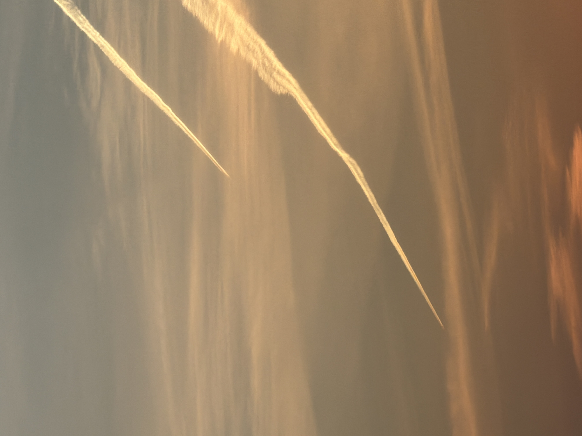 The image shows contrails in the sky. The background sky is hazy with a gradient from a light gray-blue to a warmer, peach tone. Two distinct contrails are visible, likely from airplanes. The contrails are white and dissipate slightly. Based on the location of Chaikovsky, Russia, no specific location information is available.