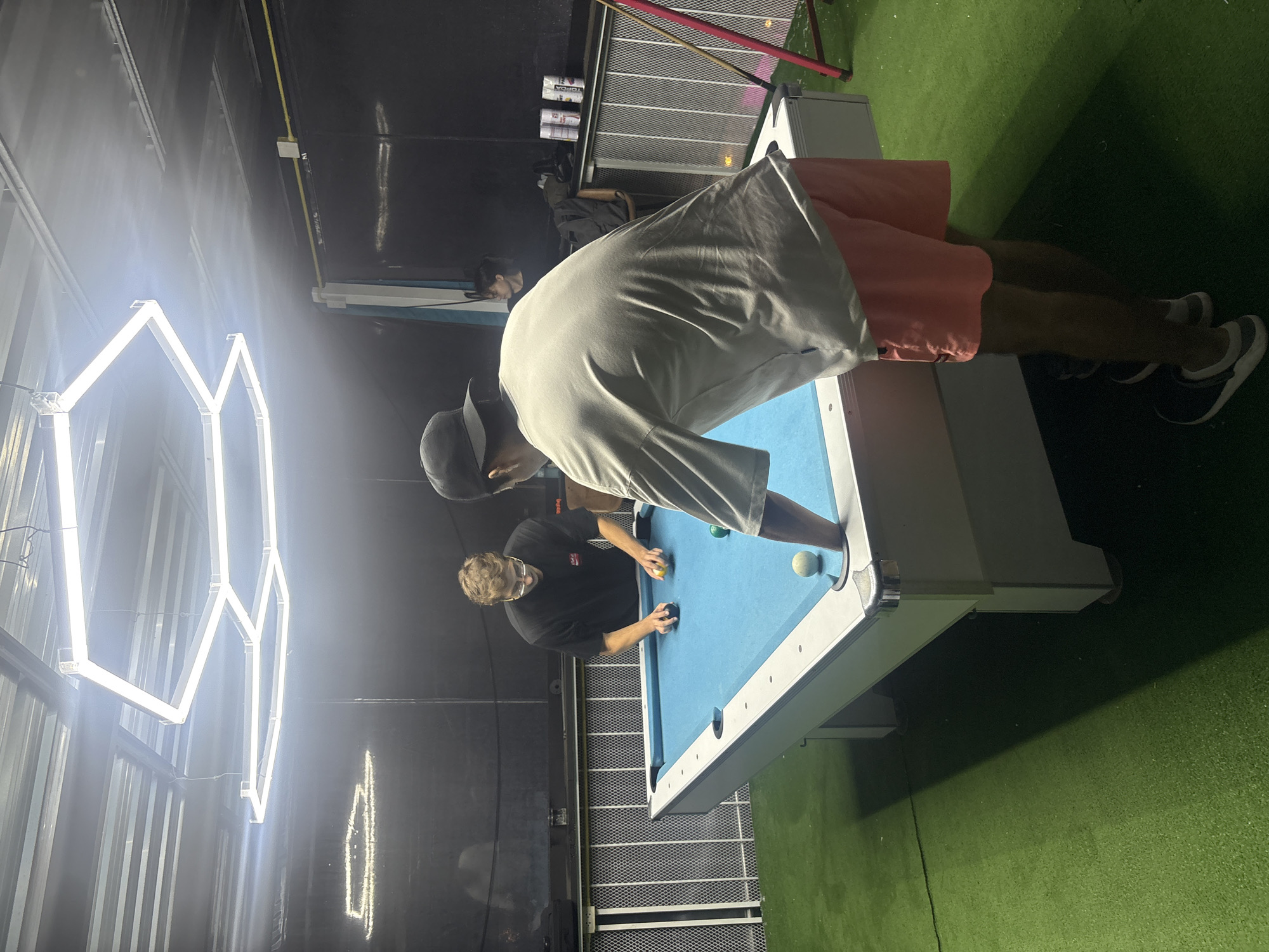 Two young men are playing pool on a blue-felt table in what appears to be an indoor recreational area. The man on the left, wearing a black t-shirt and glasses, is leaning over the table, holding a pool cue, and appears to be about to strike a ball. The man on the right, wearing a light-colored t-shirt, shorts, and a baseball cap, is standing with his back to the camera, also facing the pool table. Several pool balls are visible on the table, including a black ball, a yellow ball, a white ball, and a green ball. The floor surrounding the pool table is covered in green artificial turf. Overhead, a large, modern geometric lighting fixture illuminates the space. In the background, there is a black wall, a fence, and what looks like a bar area with some cans on it. The overall atmosphere suggests a casual leisure setting.