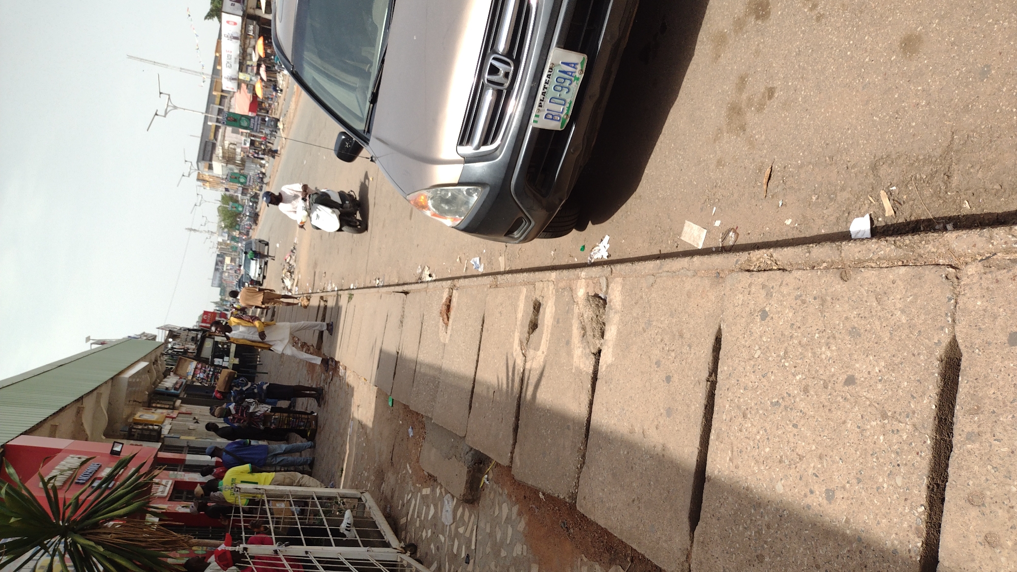 The image depicts a street scene in Jos, Nigeria. A tan-colored Honda is parked along the curb on the right, with its license plate partially visible. People are gathered on the sidewalk next to a row of shops, with one person standing out in yellow. A motorcyclist is on the street. The street is littered with debris. The sidewalk includes concrete slabs that cover a drainage ditch. The lighting indicates it is daytime.