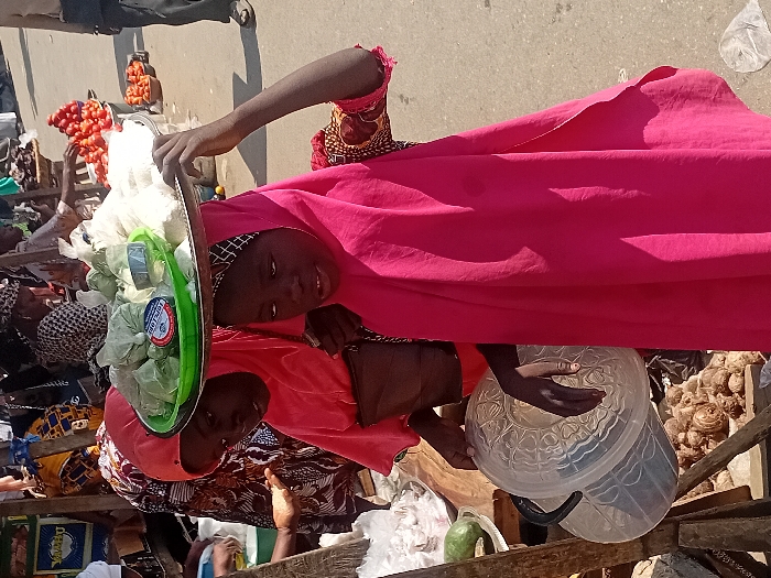 Two women at a market. One carries a tray of goods, possibly vegetables, on her head. She wears a pink headscarf. The other woman wears an orange dress with an intricate pattern, and a patterned headscarf. She holds a large plastic container. There are various goods and people in the background, creating a bustling market scene.
