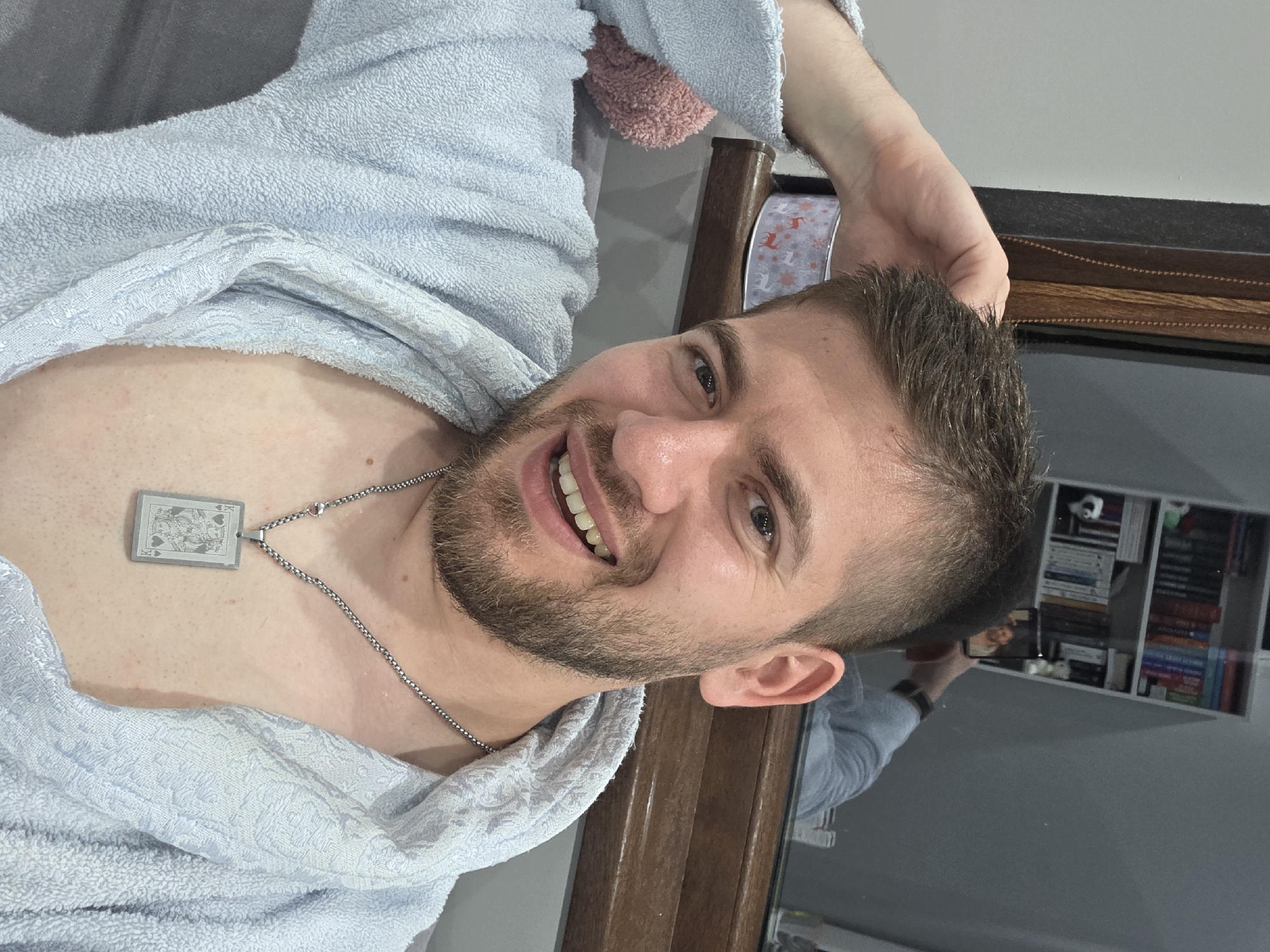 A smiling young to middle-aged man with short dark hair and stubble is featured in a close-up, rotated portrait. He looks directly at the camera with a wide, happy smile, revealing his teeth. He is wearing a light blue, textured bathrobe, which is open at the chest, showing a silver chain with a rectangular pendant. The pendant clearly depicts a King of Hearts playing card, with the letter 'K' visible. A noticeable tan line is present across his upper chest. His right arm is bent, with his hand resting near the back of his head, suggesting a relaxed pose.
The background to his right (which appears below him in the rotated image) shows a dark, reflective surface, likely a mirror, framed by dark wood. In the mirror's reflection, shelves filled with numerous books and other objects are visible. A hand holding a smartphone is also reflected, indicating that the image is a selfie. The scene is set indoors, in what appears to be a comfortable room in Yagodovo, Bulgaria. The man's expression conveys happiness and a relaxed demeanor.