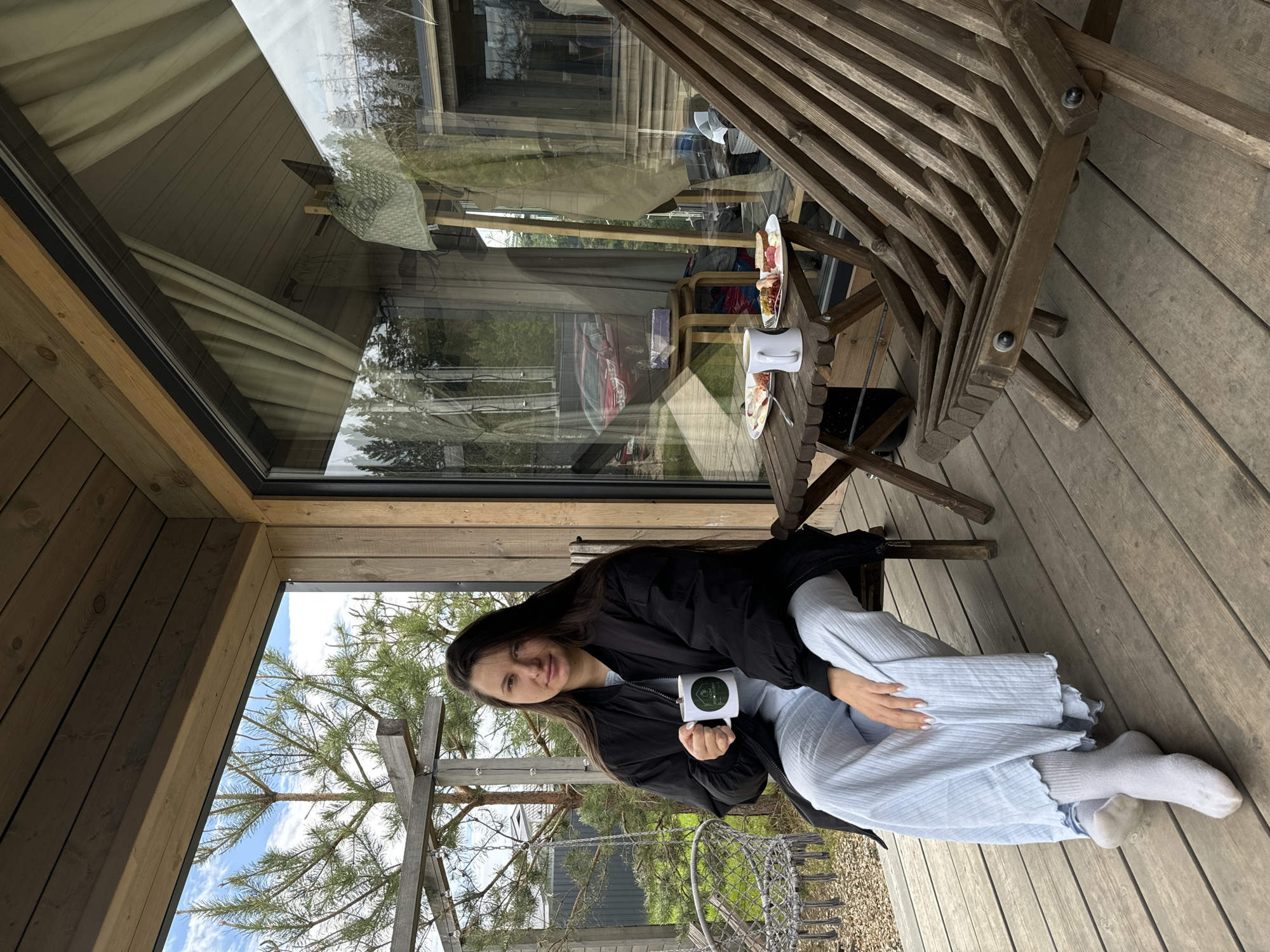 A woman is seated on a wooden deck in Литвиново, Russia, holding a white mug with a logo. She is wearing a black jacket, light blue pants, and white socks. A small wooden table with food and a white coffee pot is nearby. A wooden railing and glass windows of a building are visible in the background, as well as trees and the sky. The environment appears to be outdoors, possibly a patio or porch.