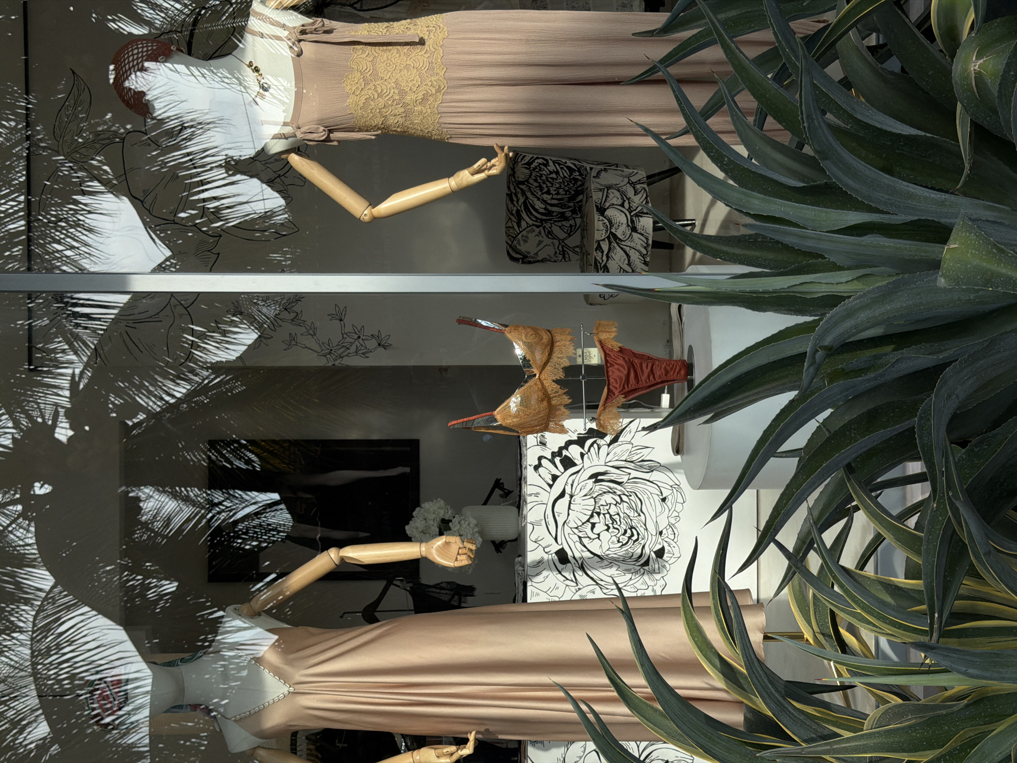 The image depicts a shop window display, reflecting the surrounding environment and obscuring the view inside. Two mannequins are visible, each dressed in a floor-length gown. The gowns appear to be made of a light-colored fabric with lace or textured detailing. The mannequins have articulated wooden arms. The background of the display features a floral design and some clothing items on hangers.
Reflections on the glass of the window add complexity to the image. The reflections include the shadow of palm trees or similar foliage. The window is also partially obscured by large agave plants.
The image is presumably taken in Mengwi, Indonesia.