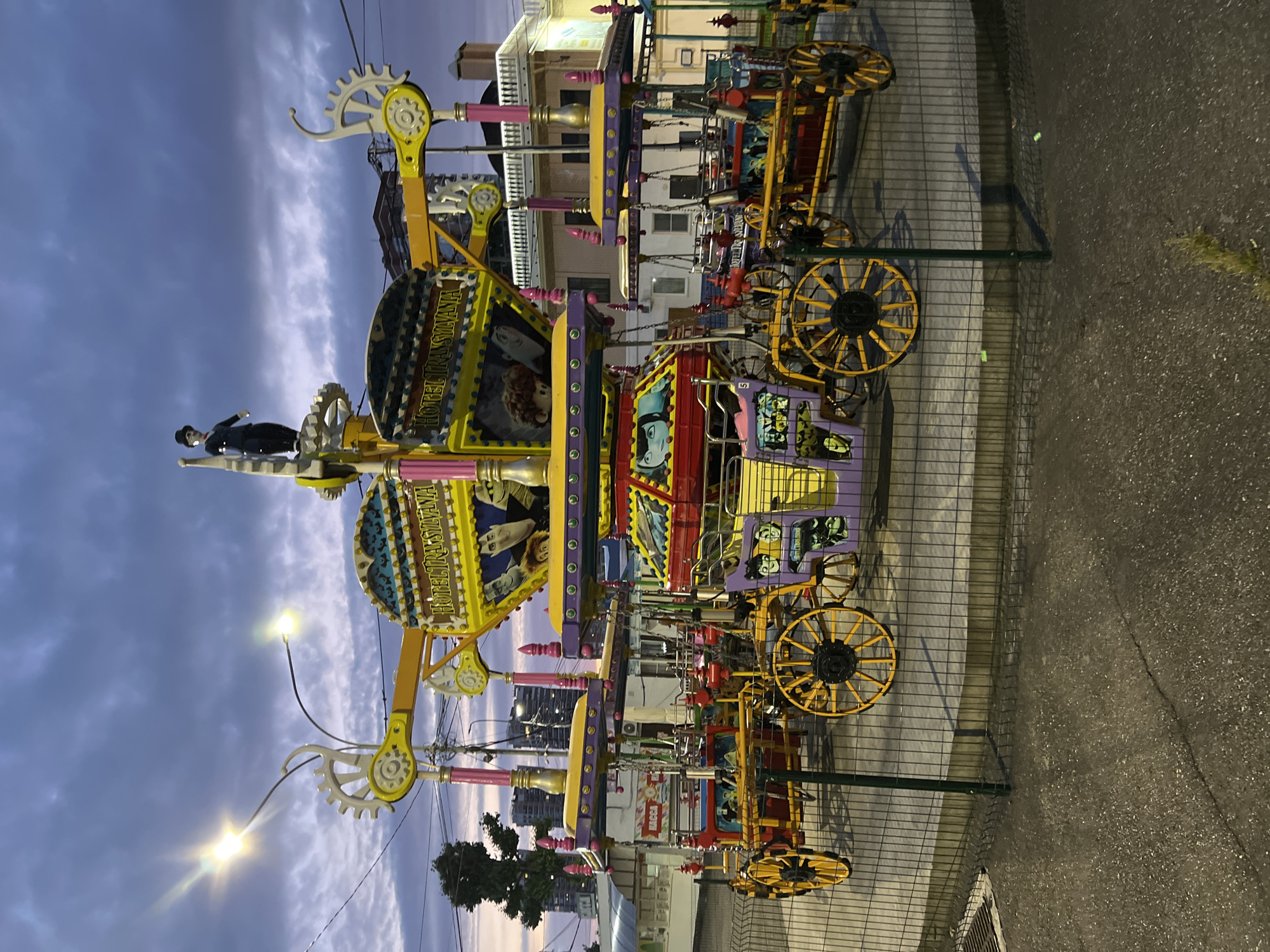 A children's amusement ride, themed after "Hotel Transylvania," is depicted at dusk in Krasnodar, Russia. The ride features colorful carriages resembling old-fashioned wagons, each with large spoked wheels. The central structure is adorned with images of characters from the "Hotel Transylvania" movie franchise, and at the very top, a figure resembling Charlie Chaplin stands. The ride is enclosed by a wire fence, and the surrounding environment includes a paved area, some greenery, and buildings in the background. The sky is overcast with dramatic clouds, illuminated by streetlights.