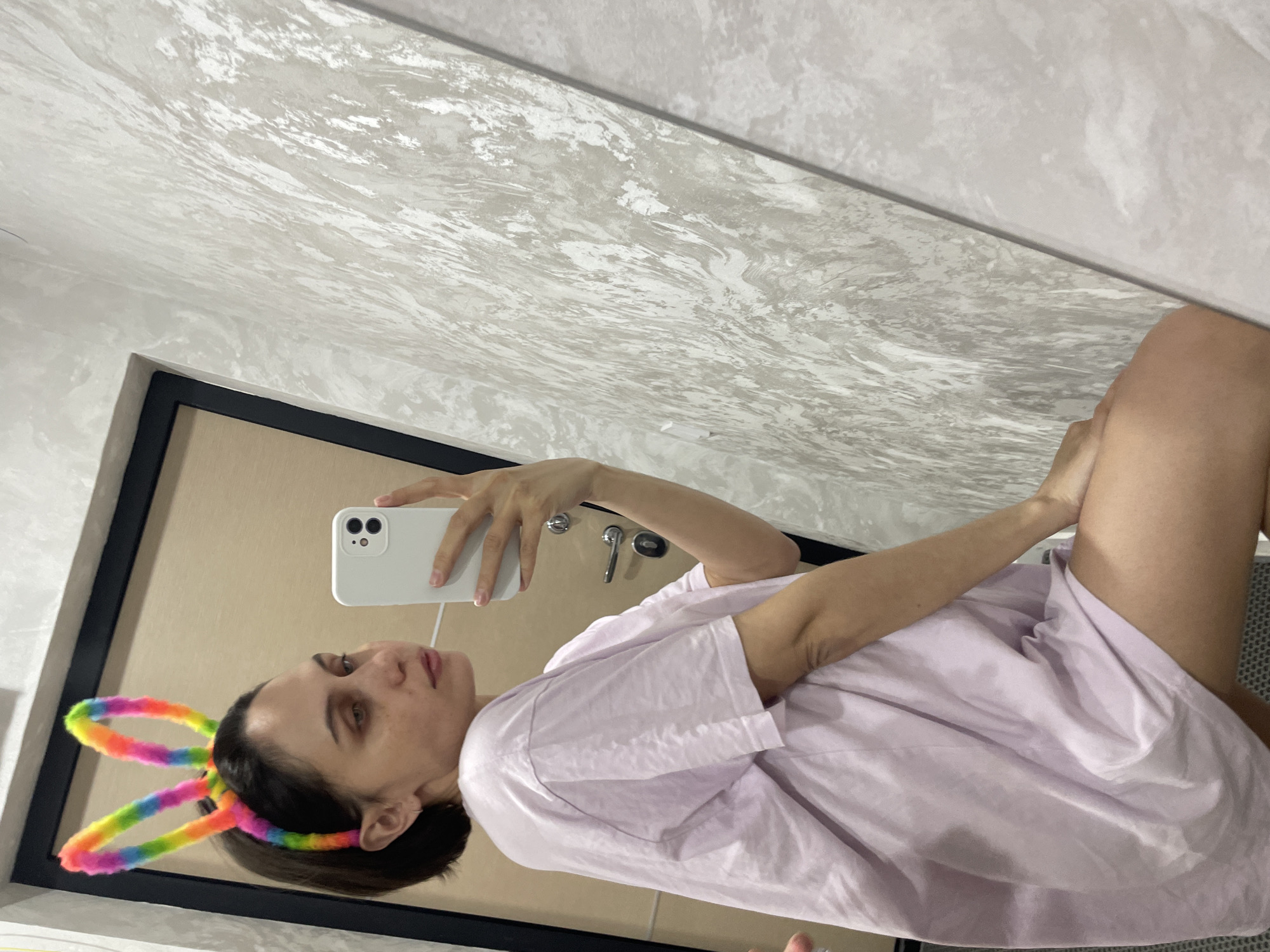 A woman with short dark hair and rainbow bunny ears takes a selfie in a bathroom mirror. She is wearing a light purple shirt. A white smartphone is held in her right hand. In the background, there is a textured wall, a doorframe, and a sink faucet visible in the mirror. There is no indication of the image being taken in Izhevsk, Russia.