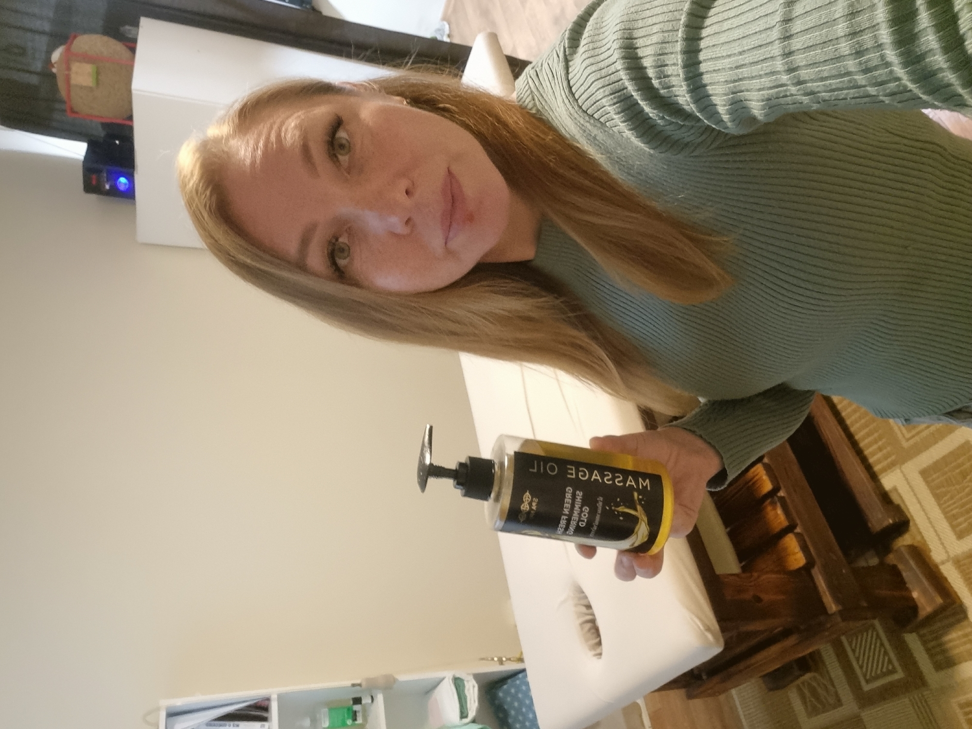 A woman with blonde hair and green eyes is holding a bottle of massage oil. The bottle has a black label with the words "Massage Oil" printed on it in gold lettering. She is wearing a green sweater. The location is a room with white walls and a massage table with a white cushion. There's a wooden structure next to the massage table. The floor has a beige carpet with a pattern. A cabinet is visible in the lower left corner with various items inside. A box is visible in the upper left corner of the image. There is no information to confirm this photo was taken in Chaikovsky, Russia.