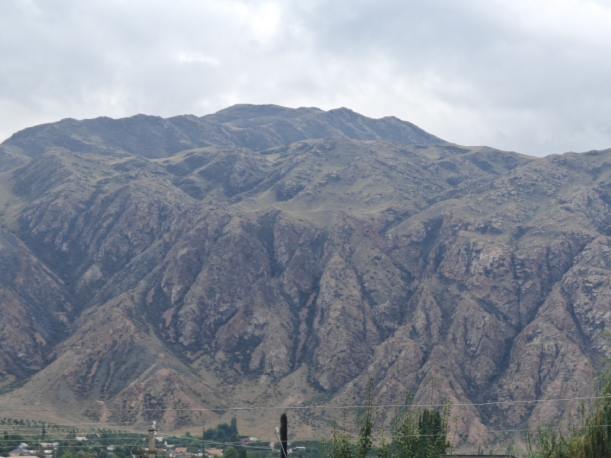 The image displays a panoramic view of rugged, barren mountains under a cloudy sky. The mountains exhibit distinct geological formations with steep slopes and visible striations. In the foreground, a village is nestled at the base of the mountains, characterized by scattered buildings, trees, and utility poles. A power line is visible spanning across the lower part of the image, running parallel to the ground. The overall scene depicts a remote, mountainous landscape adjacent to a settlement. The location is identified as Kaji-Sai 2, Kyrgyzstan.